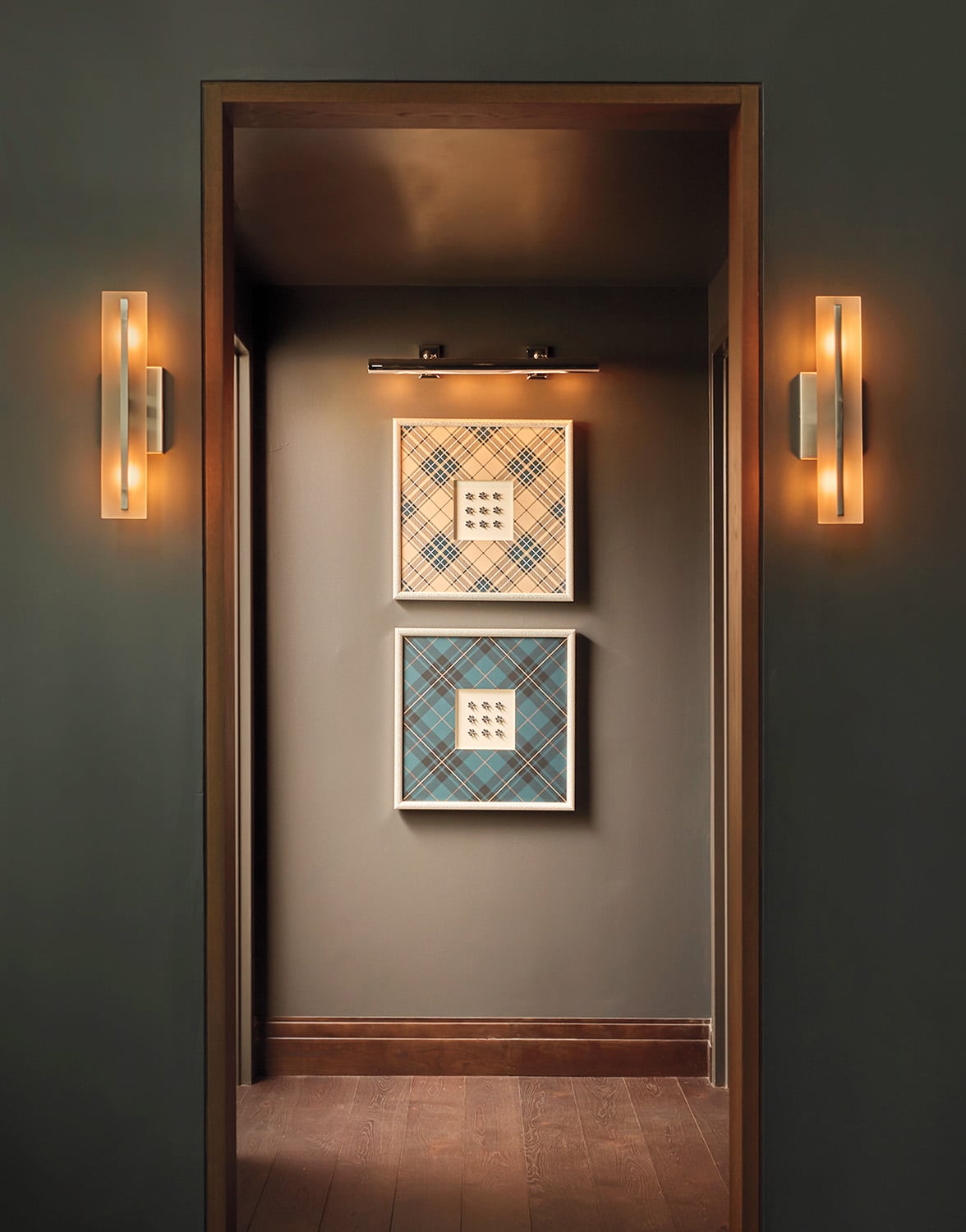 A hallway with dark walls, wooden floors, two wall sconces, and two framed geometric artworks hanging on the far wall under a picture light.