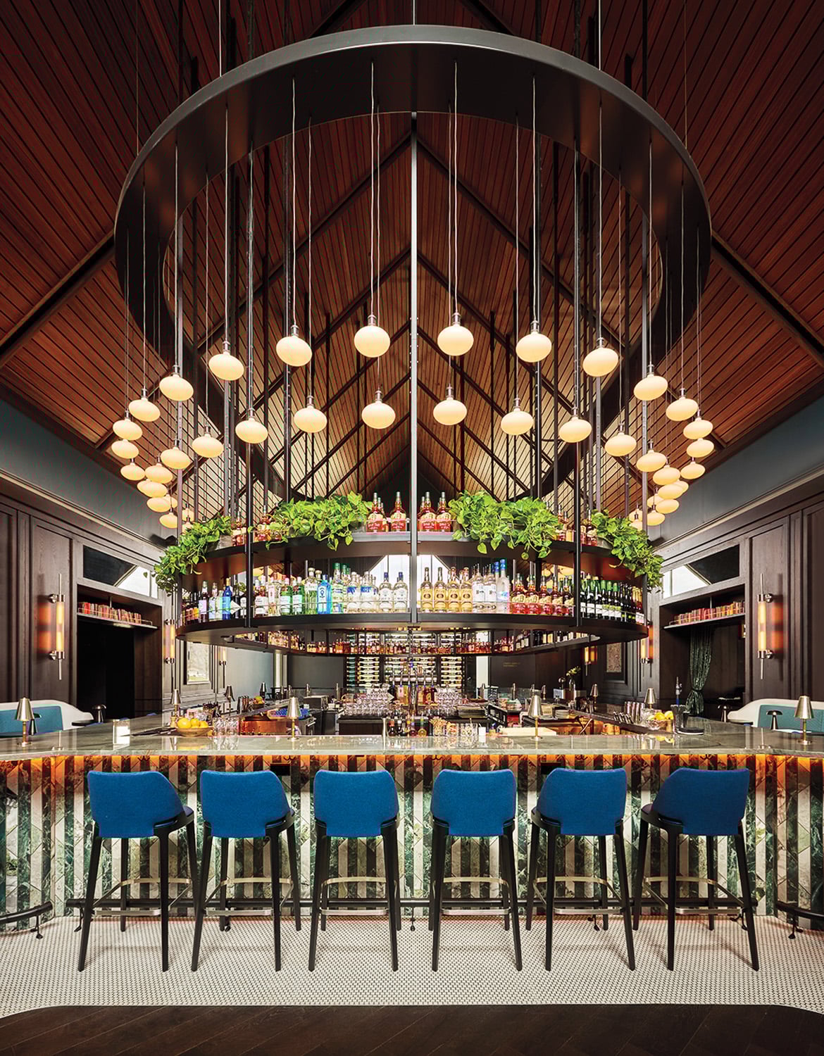 A modern bar with a high ceiling, pendant lights, a curved shelf stocked with bottles, greenery, and six blue chairs at a marble counter.