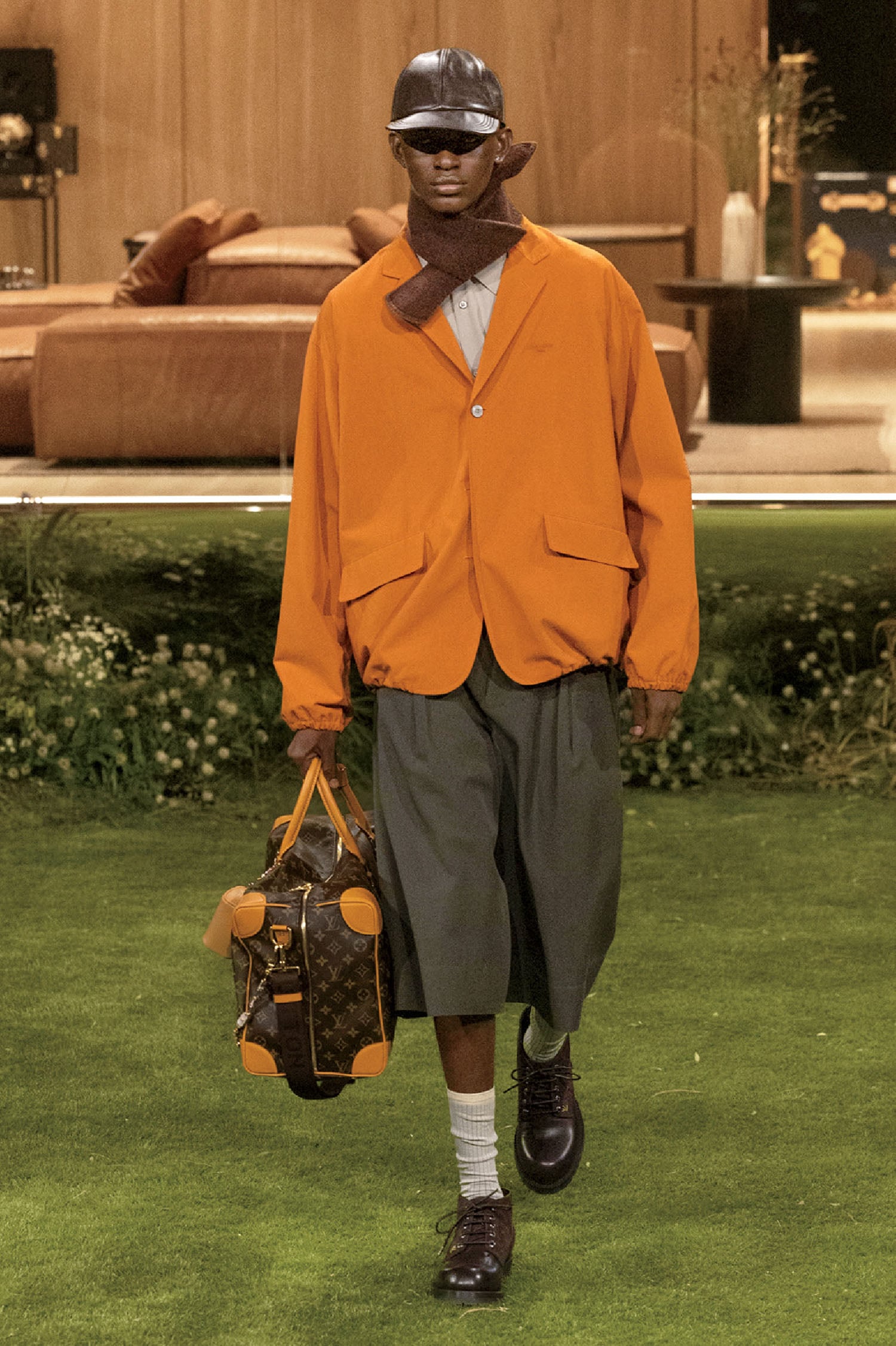 A man in a hat and orange jacket walking on a green lawn.