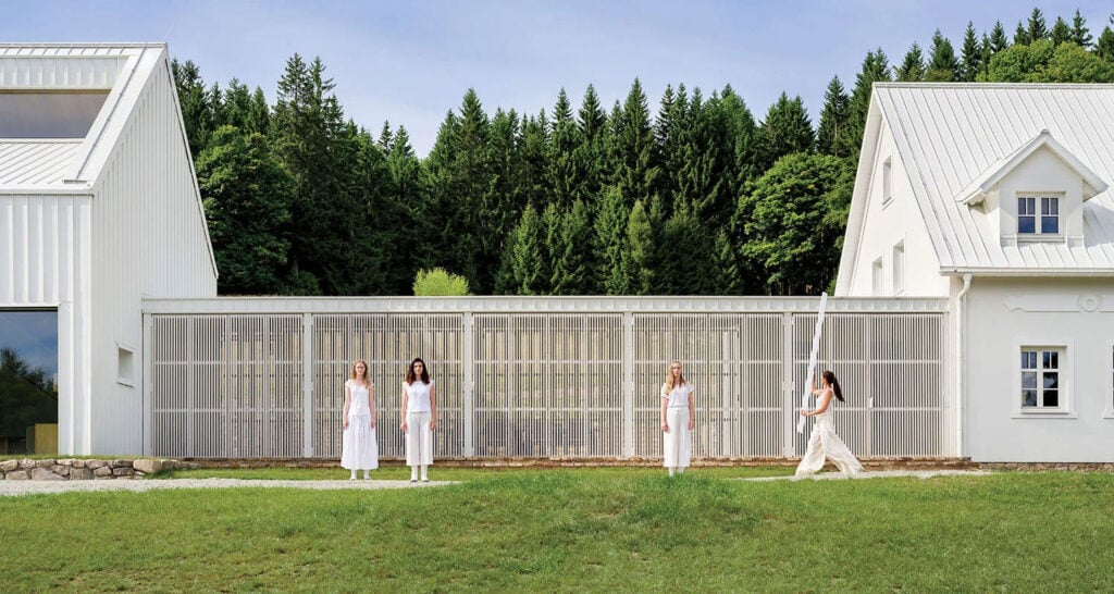 Four women in white clothing stand in front of a modern white building with a forest in the background, under a clear sky.