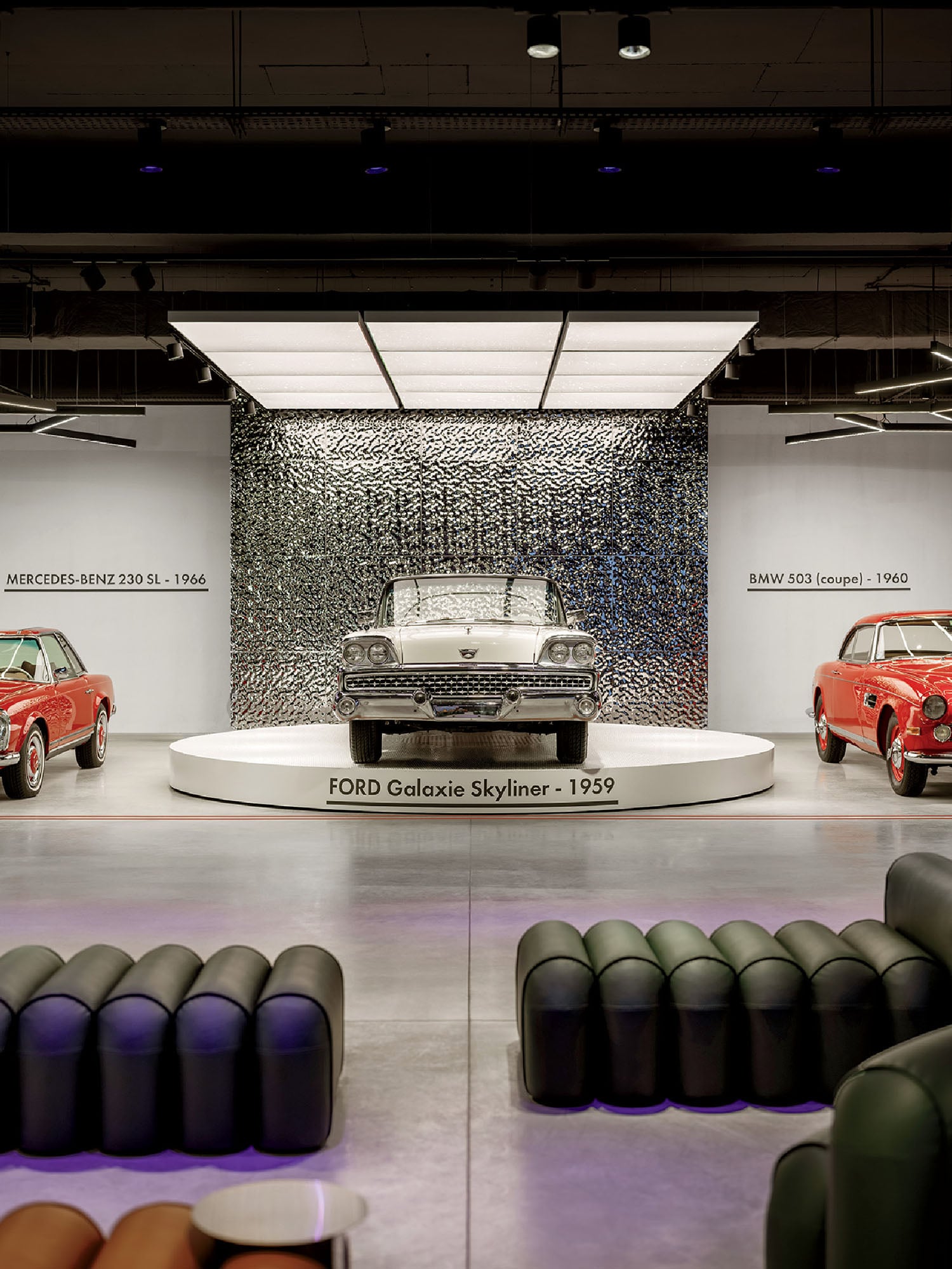 A 1959 Ford Galaxie Skyliner is displayed on a raised platform between a 1966 Mercedes-Benz 230 SL and a 1960 BMW 503 coupe in a modern indoor car exhibit.
