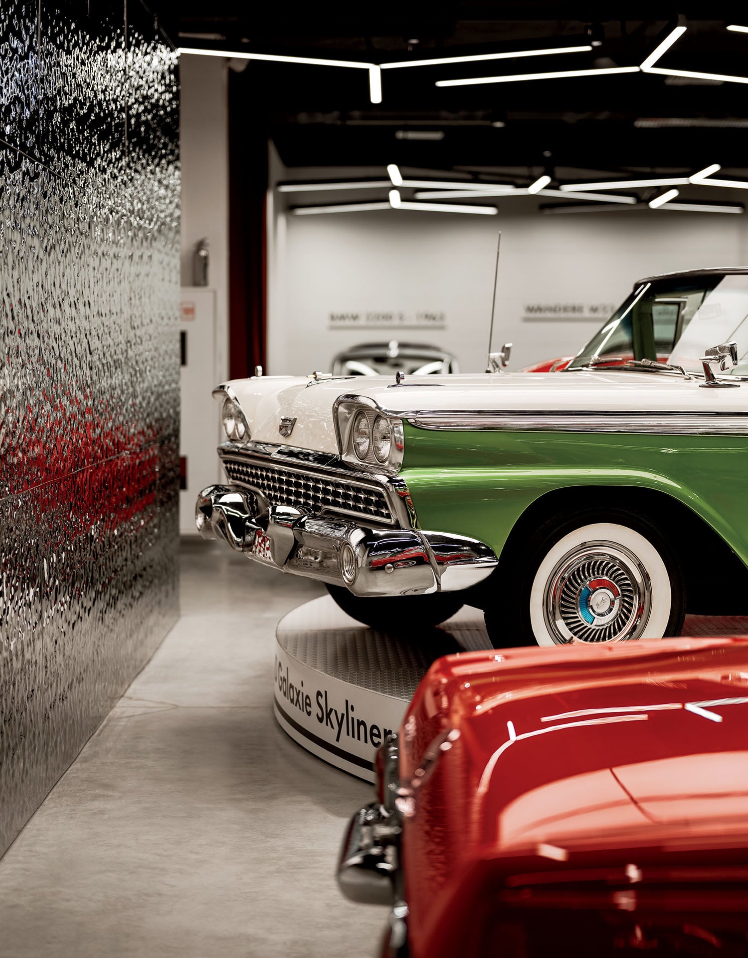 A green and white vintage Ford Fairlane 500 Skyliner convertible displayed on a platform beside a red classic car in an indoor car museum.