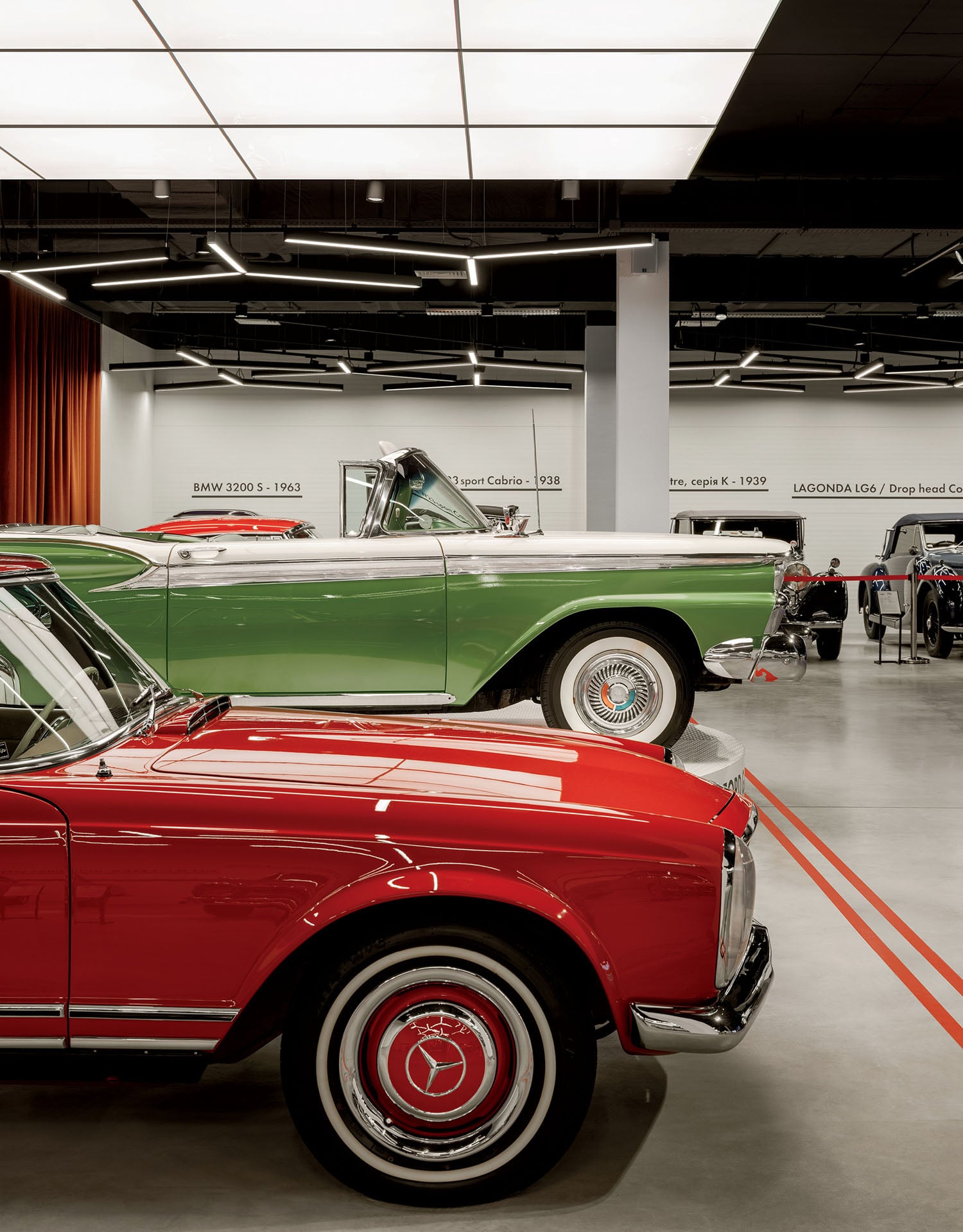 Two classic cars, one red and one green convertible, are displayed indoors under bright ceiling lights in a modern showroom.