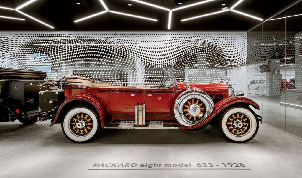 A red Packard Eight Model 633 car from 1928 is displayed in a modern showroom with a patterned metal backdrop and overhead lighting.