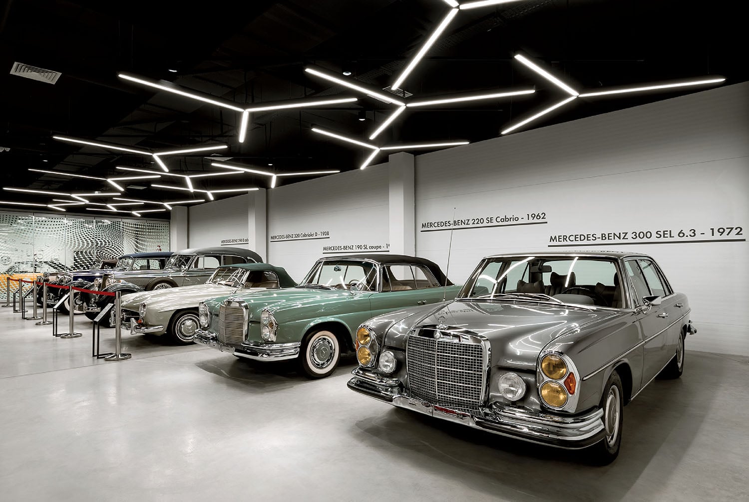 A row of classic Mercedes-Benz cars displayed in a modern showroom with geometric ceiling lights and information on the wall behind each vehicle.