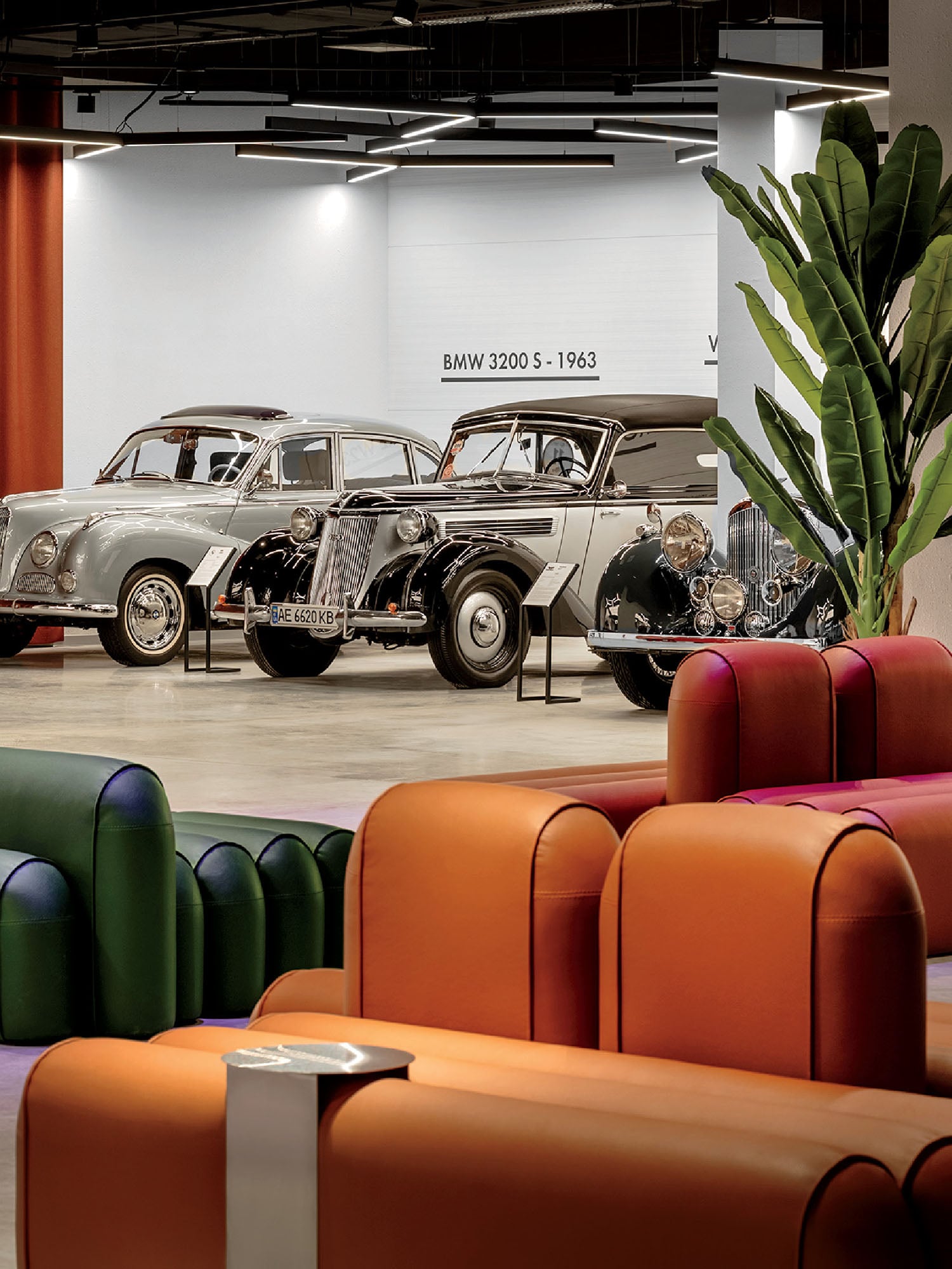 Three vintage cars are displayed in a modern showroom with colorful seating and a large indoor plant; the wall behind reads “BMW 3200 S - 1963.”.