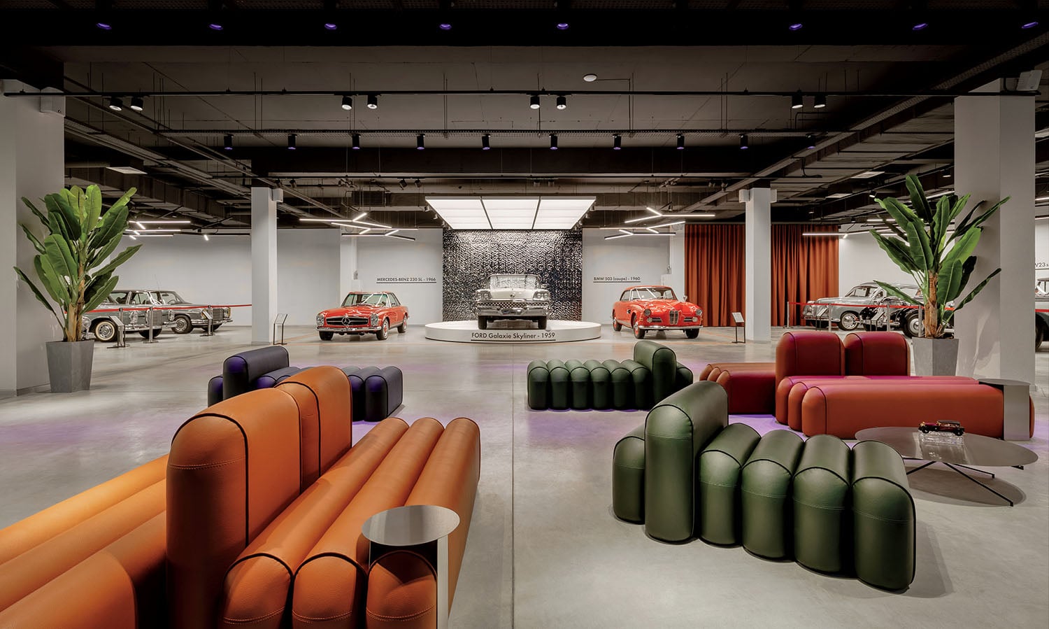 Modern showroom with vintage cars on display, colorful modular sofas, plants, and industrial-style ceiling lighting.