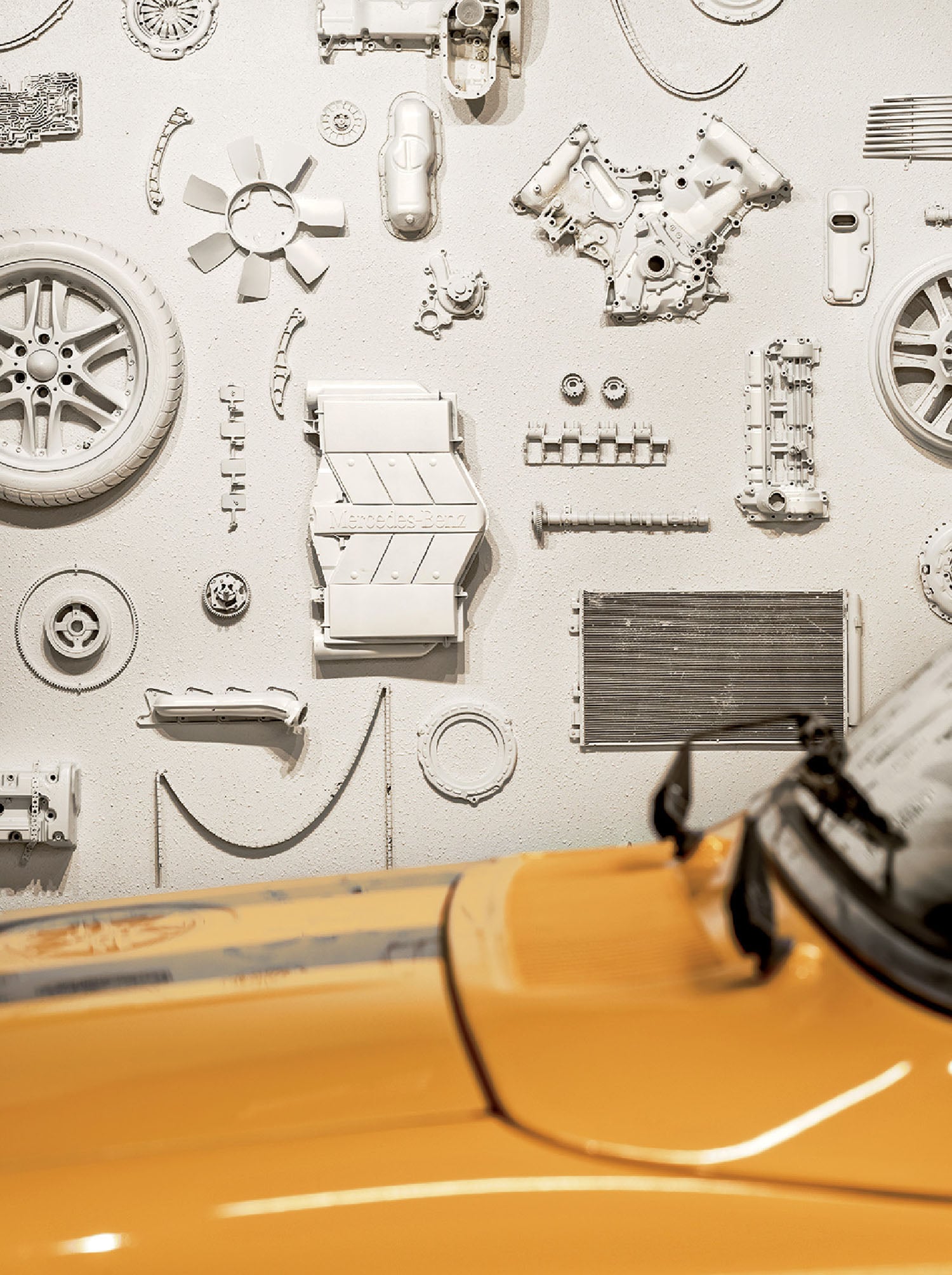 A yellow car is parked in front of a wall displaying neatly arranged, white-painted car parts and mechanical components.