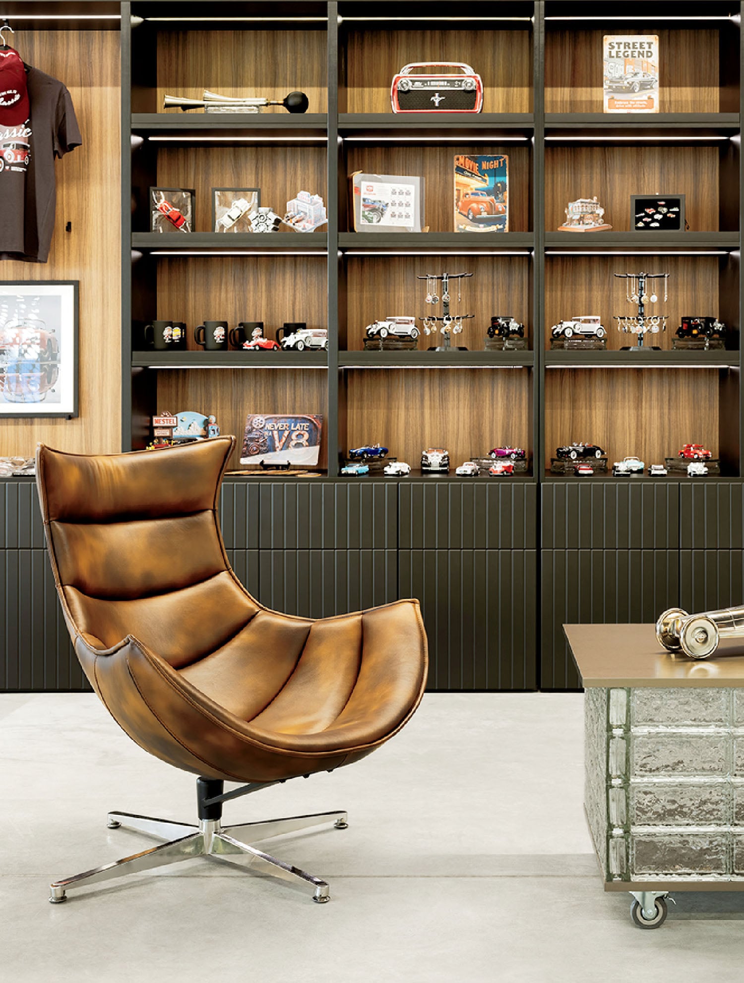 A brown leather swivel chair sits in front of a bookshelf displaying model cars, car memorabilia, and collectibles, with a glass coffee table and headphones nearby.