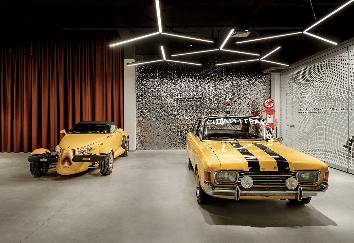 Two yellow cars are displayed in a modern showroom with geometric ceiling lights and a metallic textured wall in the background.