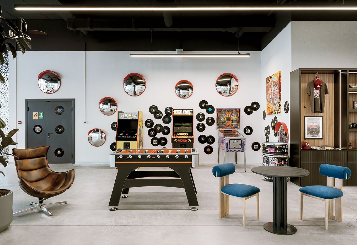 Modern recreation room with foosball table, retro arcade machines, vinyl records and mirrors on the wall, blue chairs around a small table, and a brown leather swivel chair.