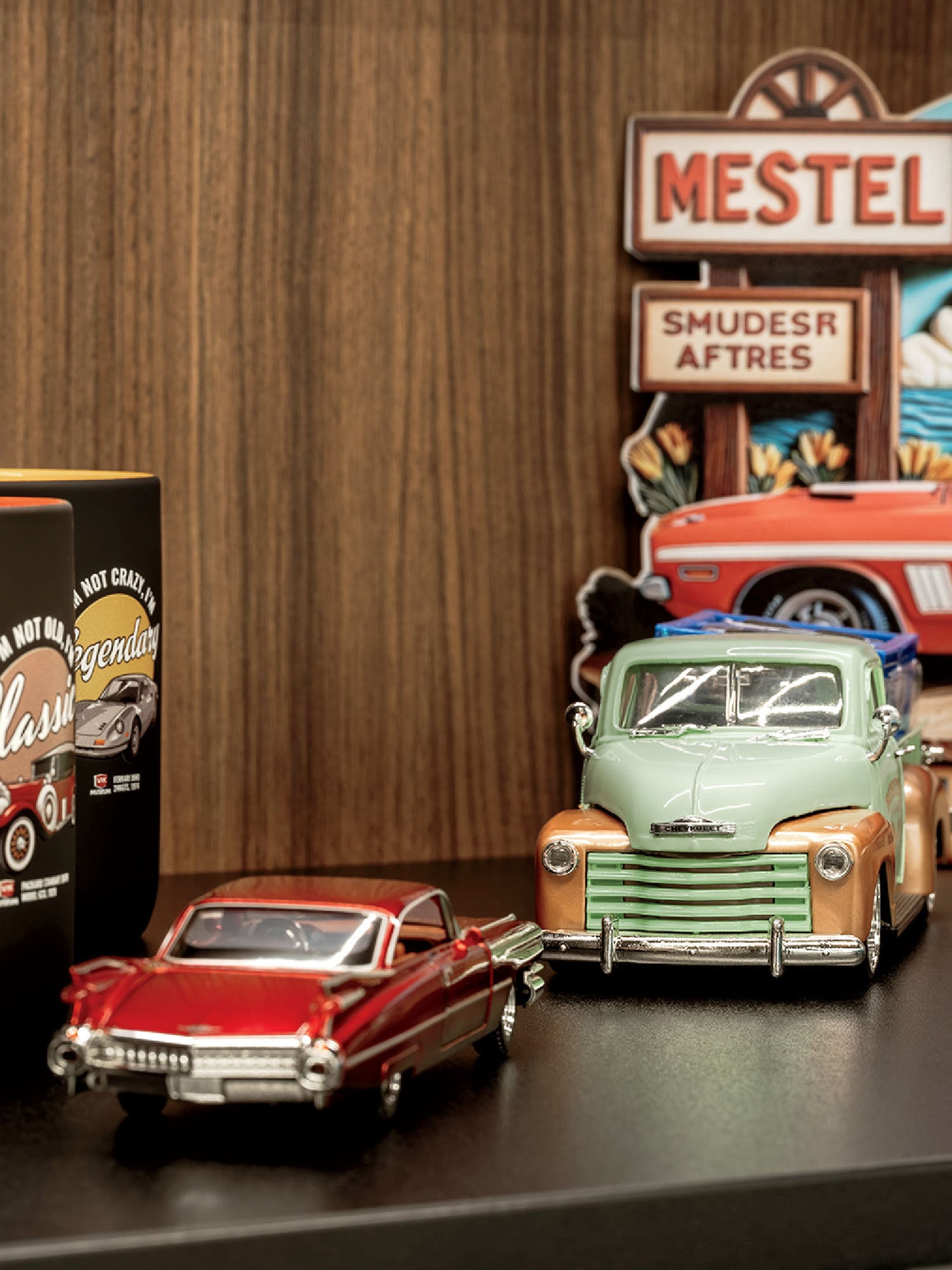 Two model cars, one red and one green, are displayed on a shelf with vintage-style automotive signs and packaging in the background against a wood panel wall.