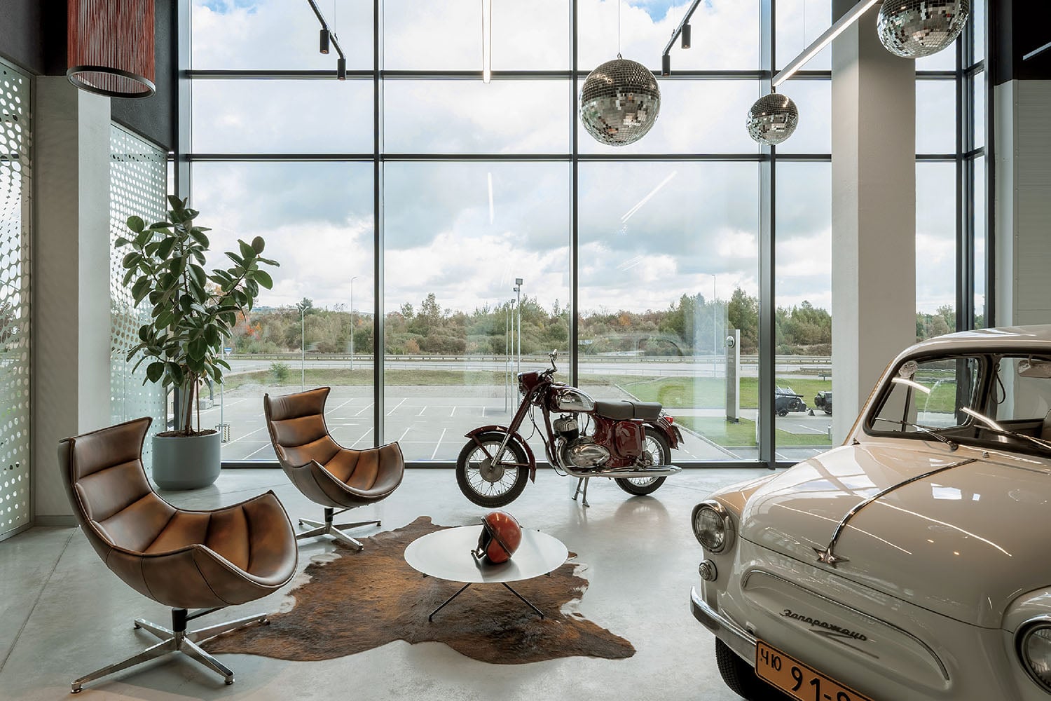Modern lounge area with leather chairs, a cowhide rug, a potted plant, a classic motorcycle, and a vintage car near large floor-to-ceiling windows, with disco balls overhead.