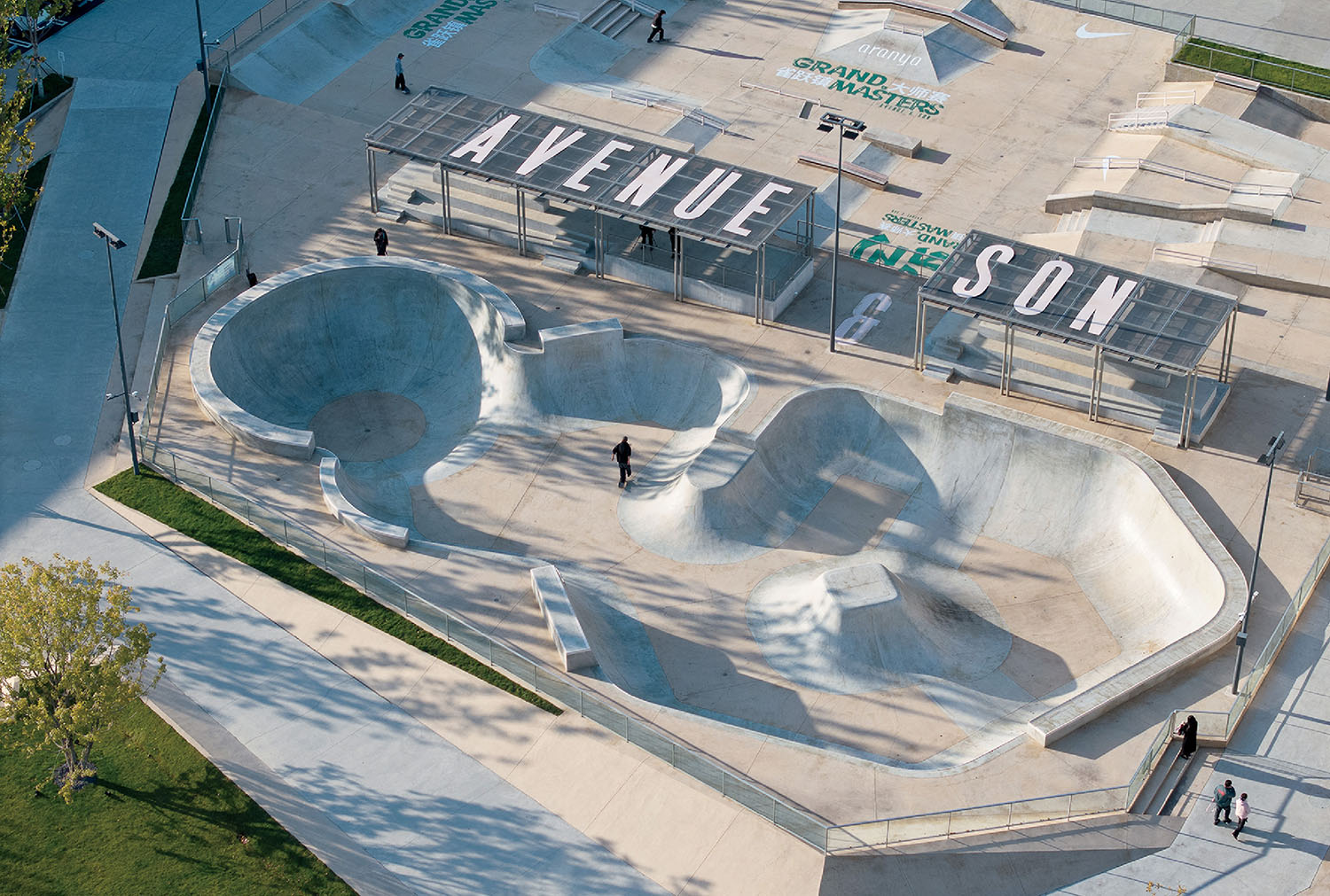 Aerial view of a concrete skatepark with multiple bowls, ramps, and a shaded area labeled "AVENUE & SON." A few people are skating and walking in the park.