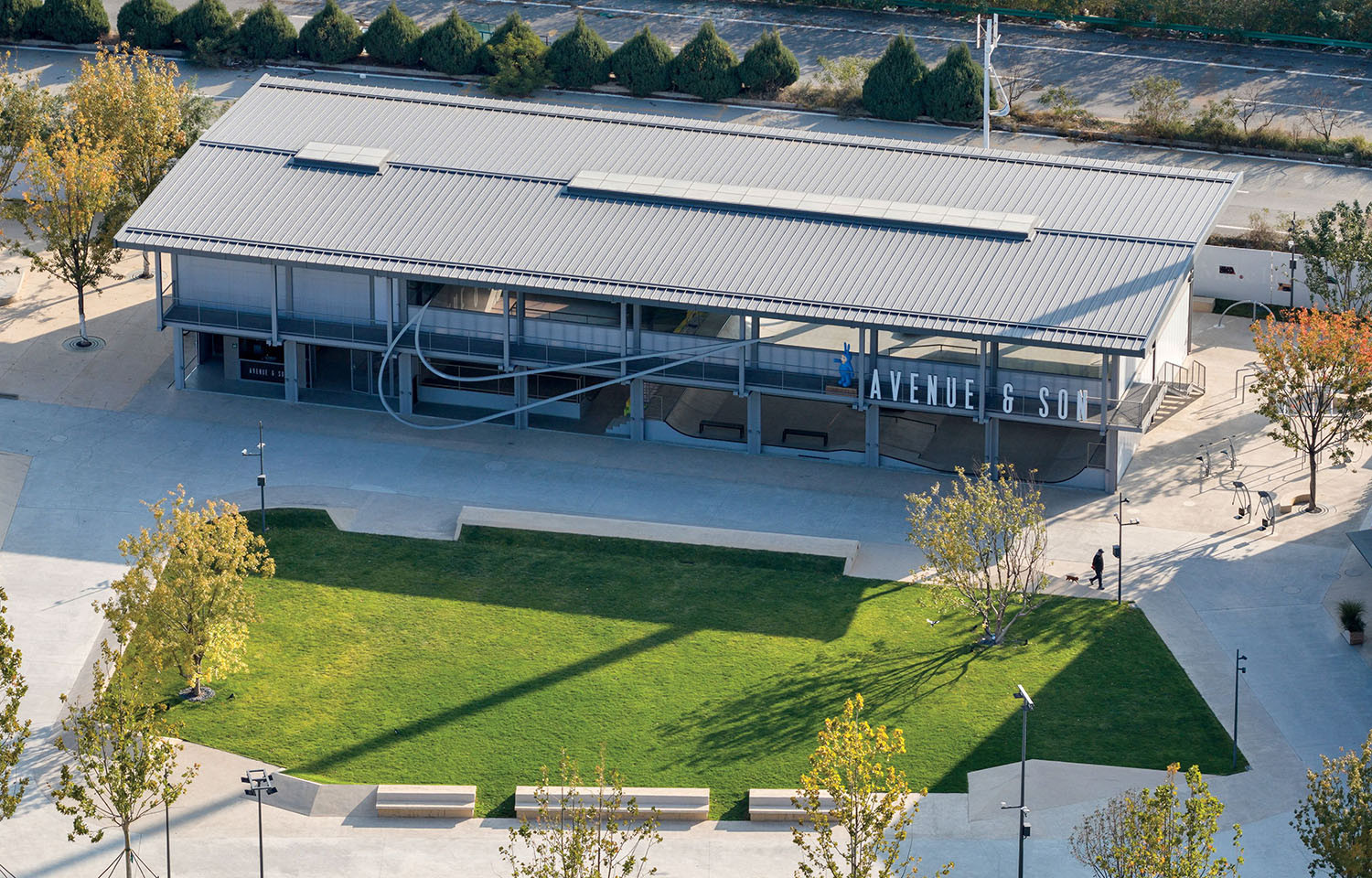 Aerial view of a modern two-story building with "Avenue & Sol" sign, surrounded by a green lawn, trees, and walkways in an outdoor plaza setting.