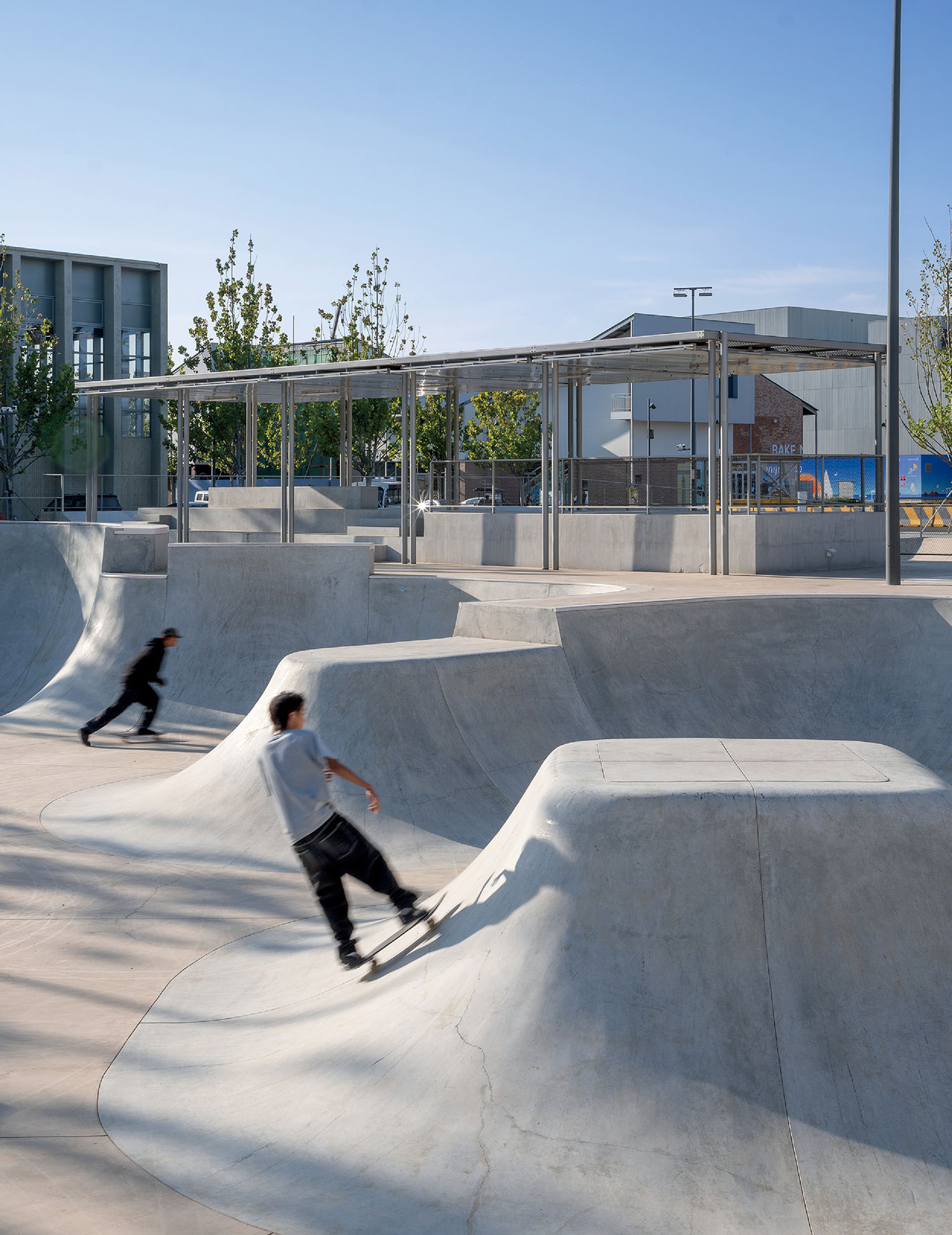 Two people skateboarding in a modern concrete skatepark with ramps and curves, under clear blue sky, with buildings and trees in the background.