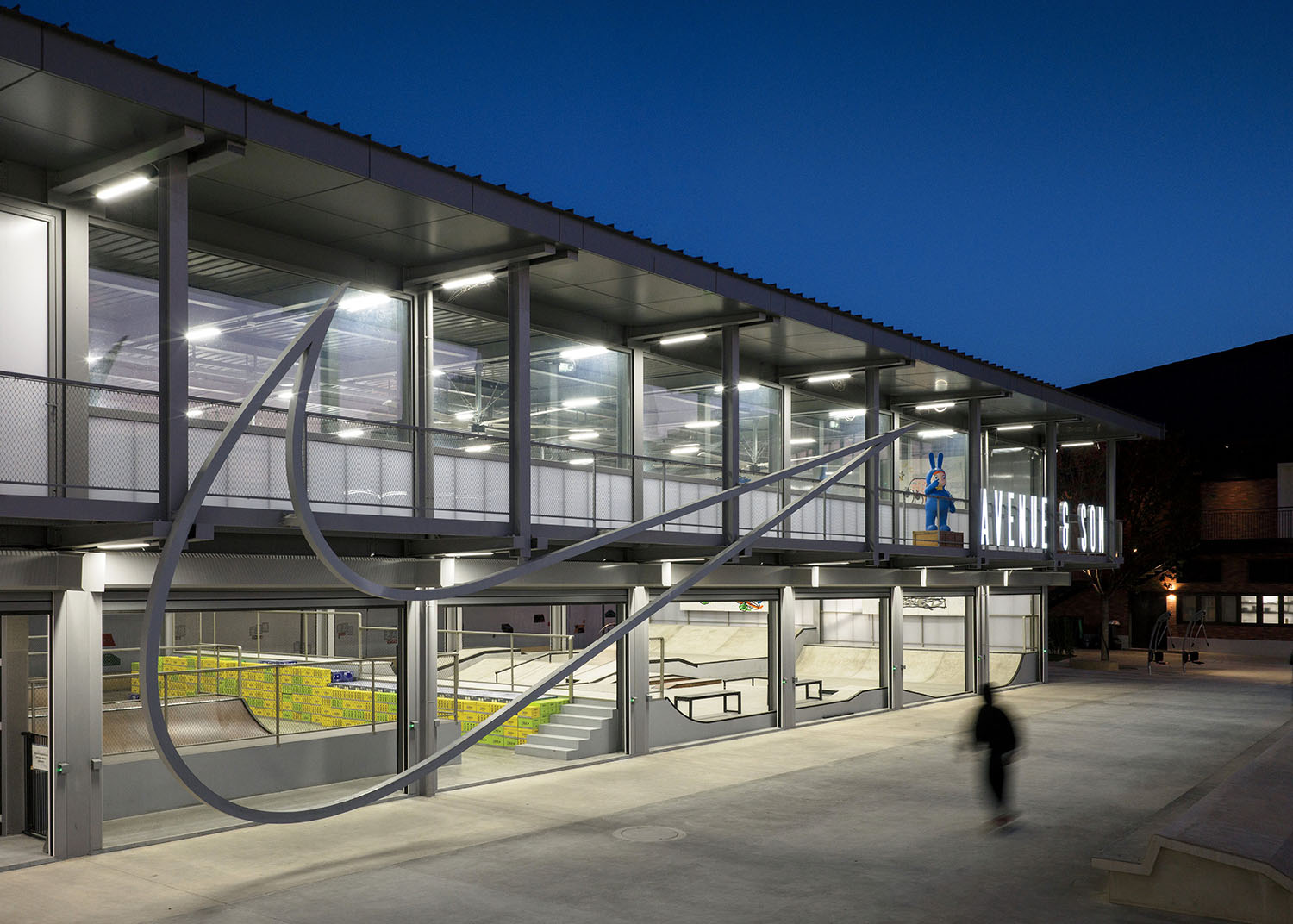 Modern two-story building at dusk with large illuminated glass windows and a prominent Nike swoosh on the facade; skateboarding ramps visible inside.