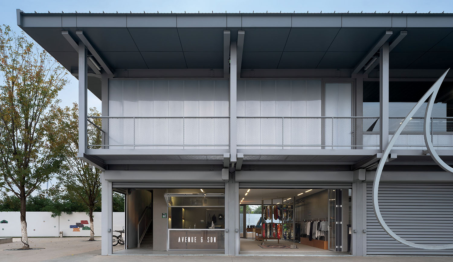 Two-story modern building with a metal facade, large windows, and an open ground floor retail space labeled "Avenue & Sun." Clothing and accessories are displayed inside.