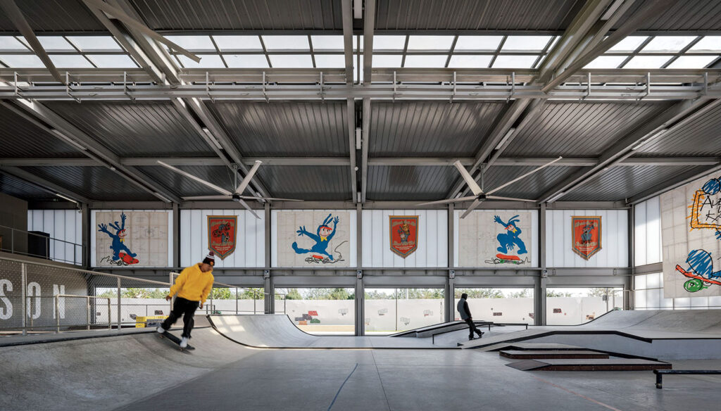 Two people skateboarding inside a spacious indoor skatepark with high ceilings, large windows, and colorful murals on the walls.