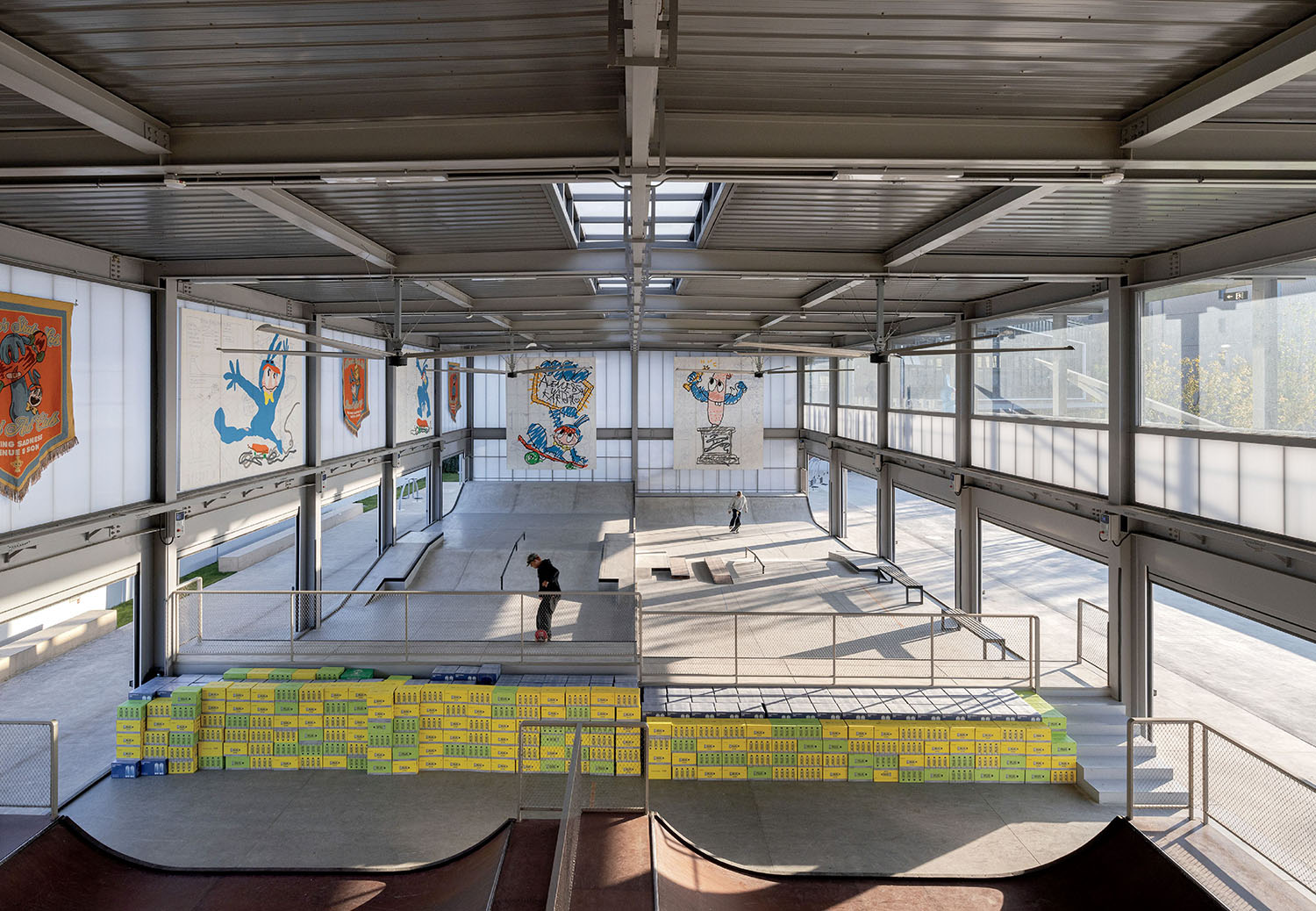 Indoor skate park with ramps, obstacles, and two people skating. Walls are decorated with colorful murals, and natural light enters through large windows and skylights.