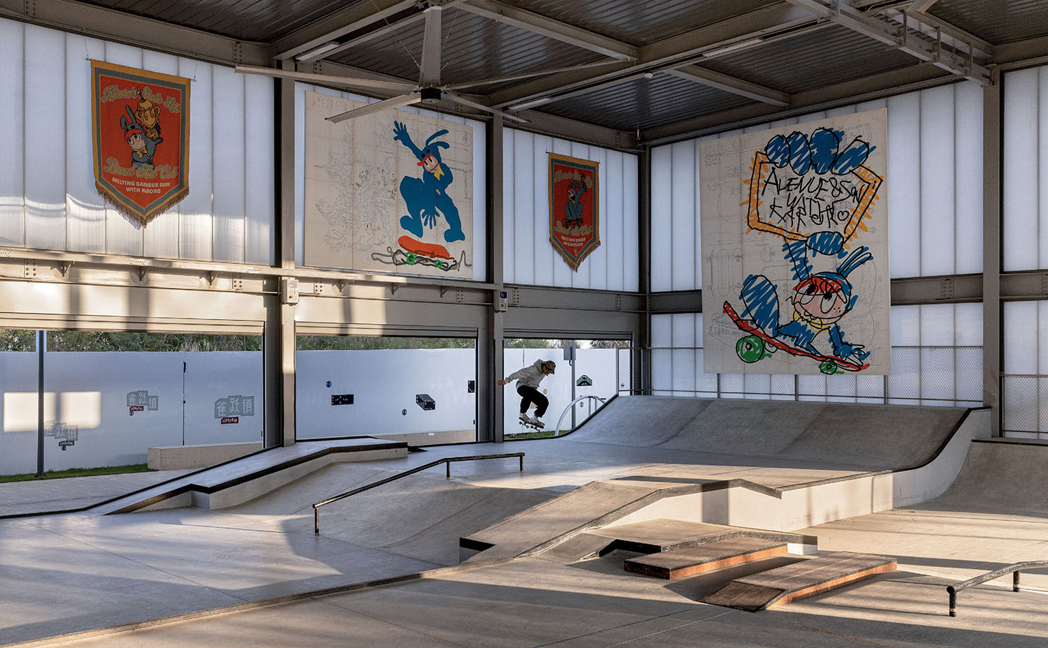 A person skateboards inside an indoor skate park with ramps and large cartoon murals on the walls.