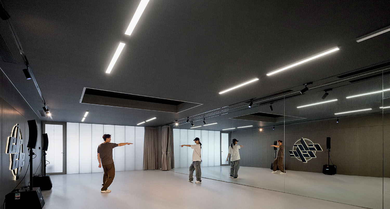 A person practices dance moves in a spacious studio with mirrored walls, LED ceiling lights, and a minimalist interior.