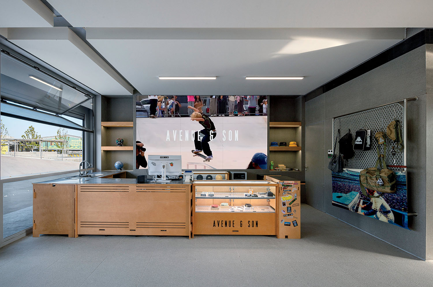 Skate shop interior with a wooden counter, display case, skateboard accessories, and a large screen showing a skateboarder with "Avenue & Son" text in the background.