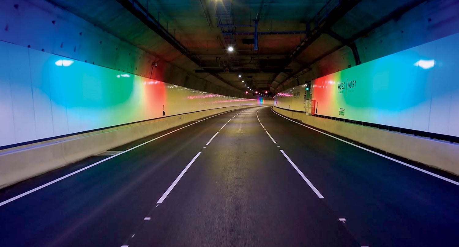 A wide, empty tunnel with reflective white walls illuminated by colored lights and three lanes separated by a solid barrier.