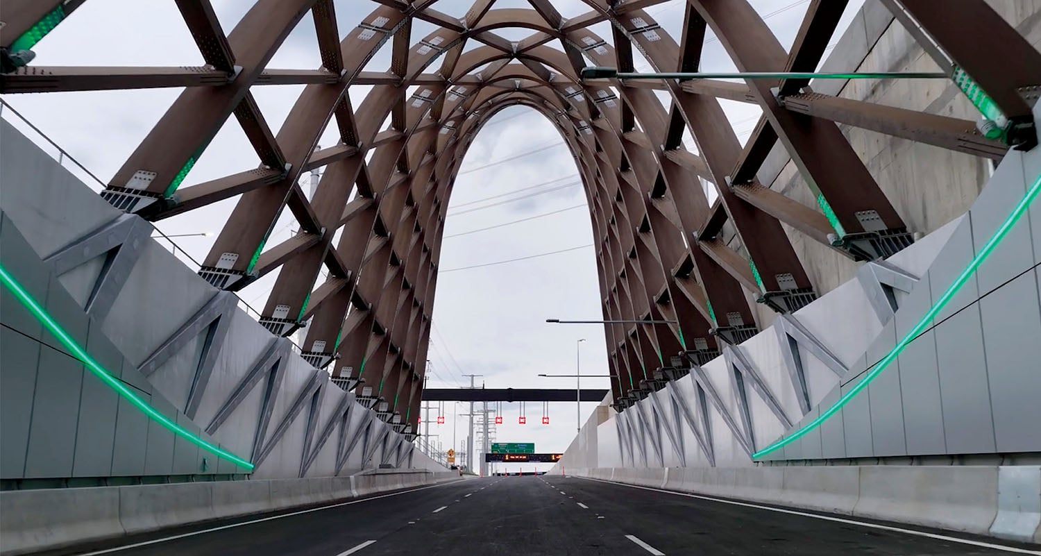 View from the road inside a modern tunnel with a geometric lattice ceiling and green accent lighting along the concrete walls.