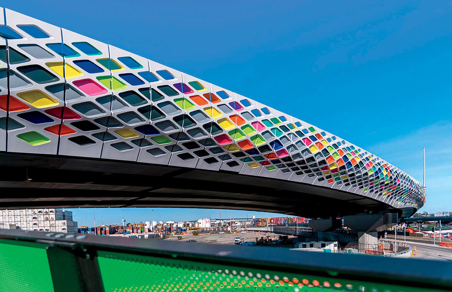 Curved bridge with a facade of diamond-shaped cutouts, some filled with colorful panels, spans over an industrial area under a clear blue sky.
