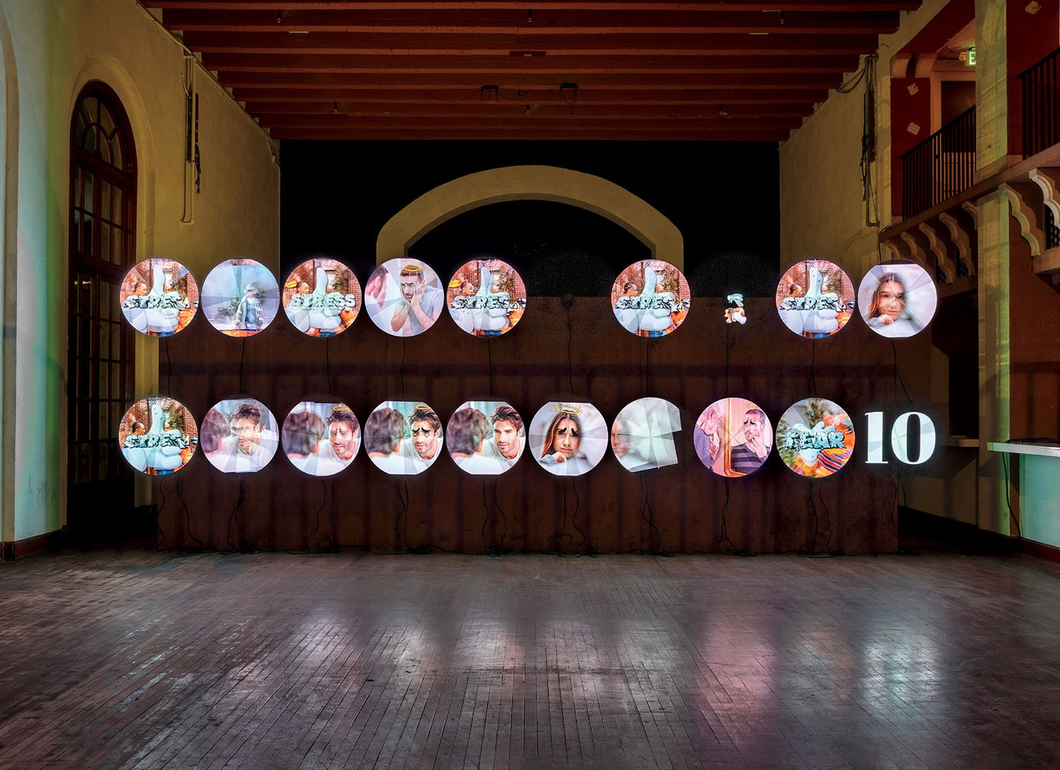 A row of circular screens displays various faces and scenes above a wooden structure in a dimly lit, spacious room with arched windows and decorative walls.