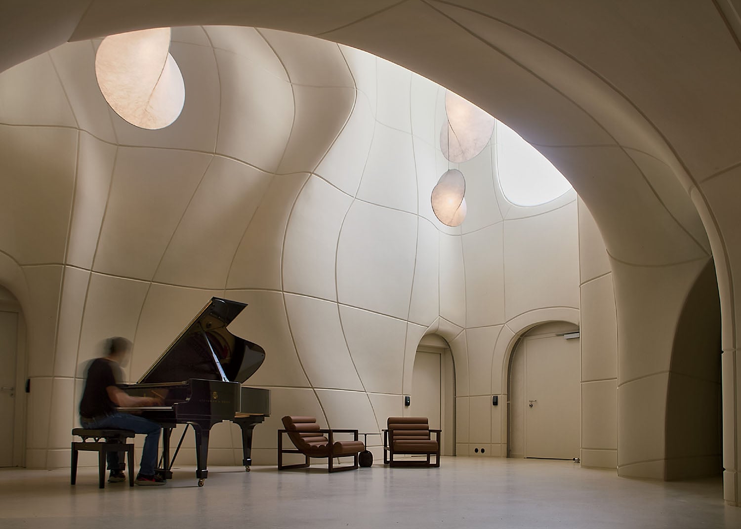 A person plays a grand piano in a spacious room with curved, padded walls, circular skylights, and minimalist furniture.