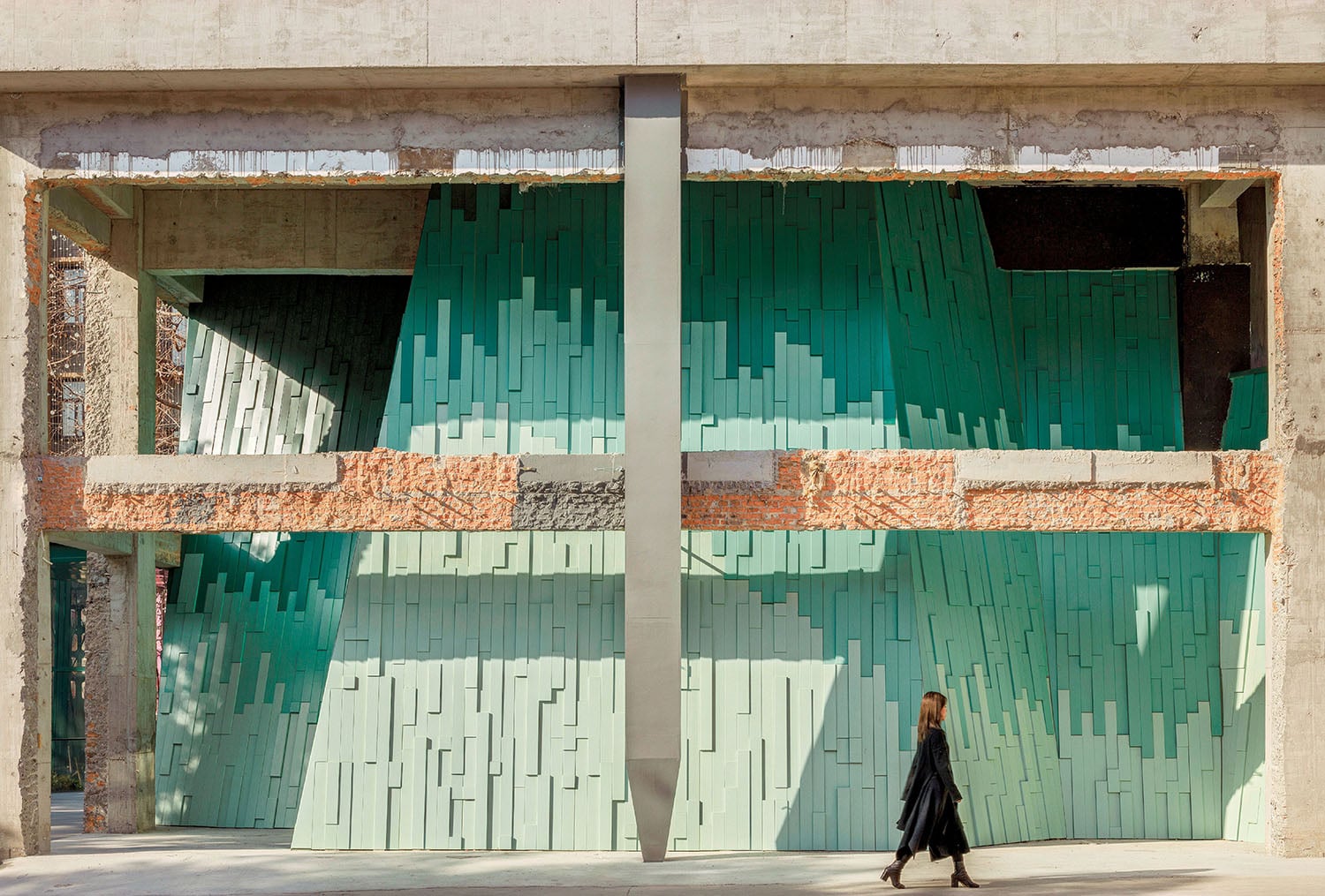 A person walks past a building facade with exposed bricks and teal, vertically stacked panels forming an abstract geometric pattern.