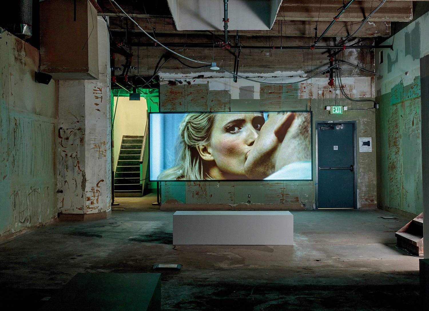 A large screen in an industrial room displays a close-up of a woman kissing a man's face. A white bench sits in front of the screen; a door and stairs are in the background.