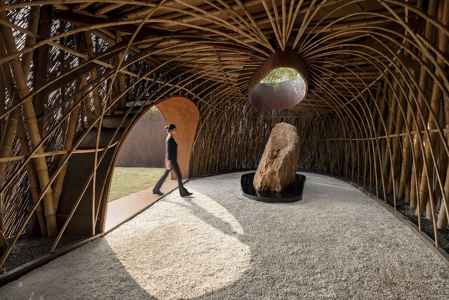 A person walks through a bamboo-structured pavilion with a large rock centerpiece and gravel floor, illuminated by natural light through curved openings.
