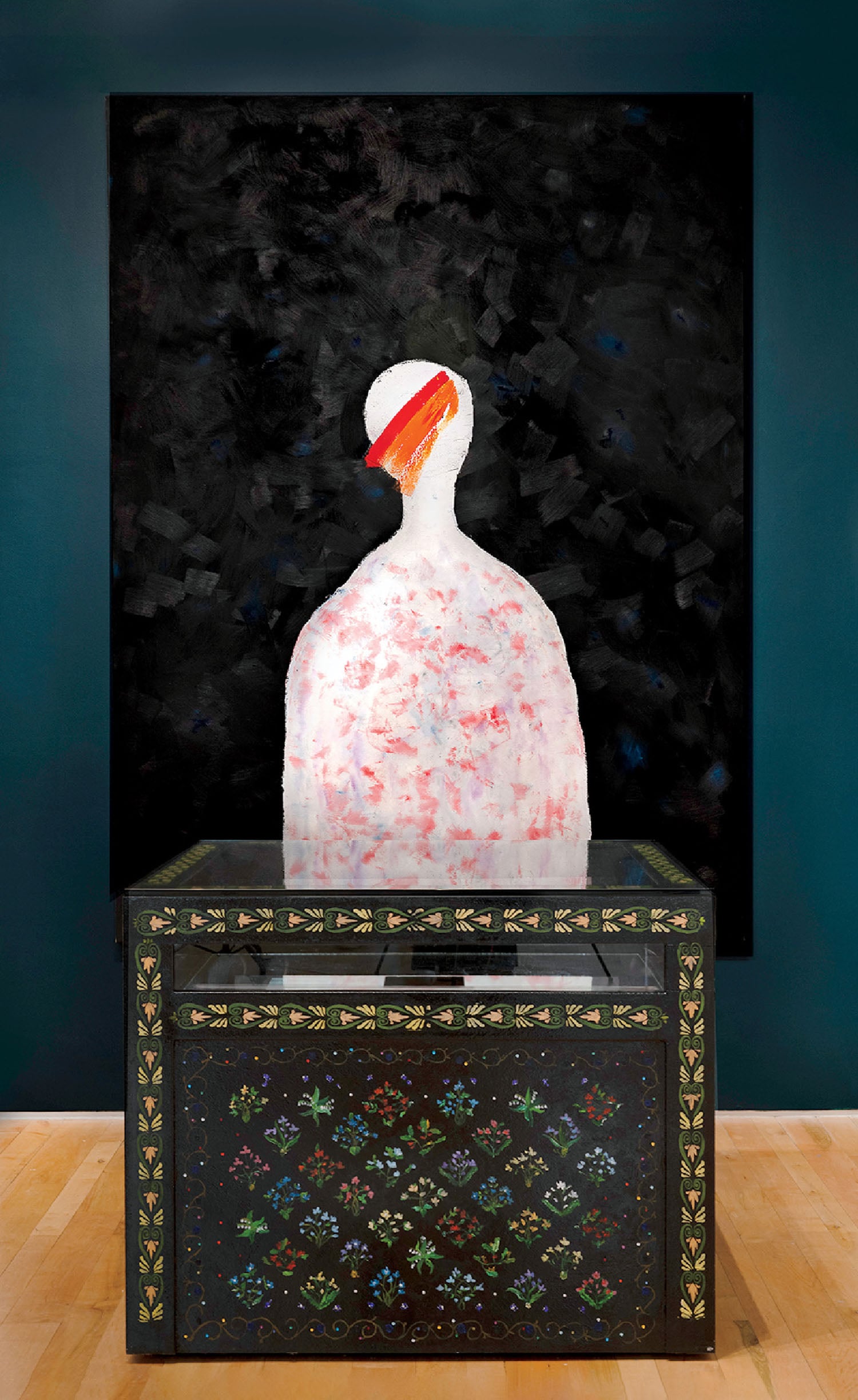 A patterned chest sits in front of a painting depicting a white, abstract human figure with a red and orange streak across its head on a black background.