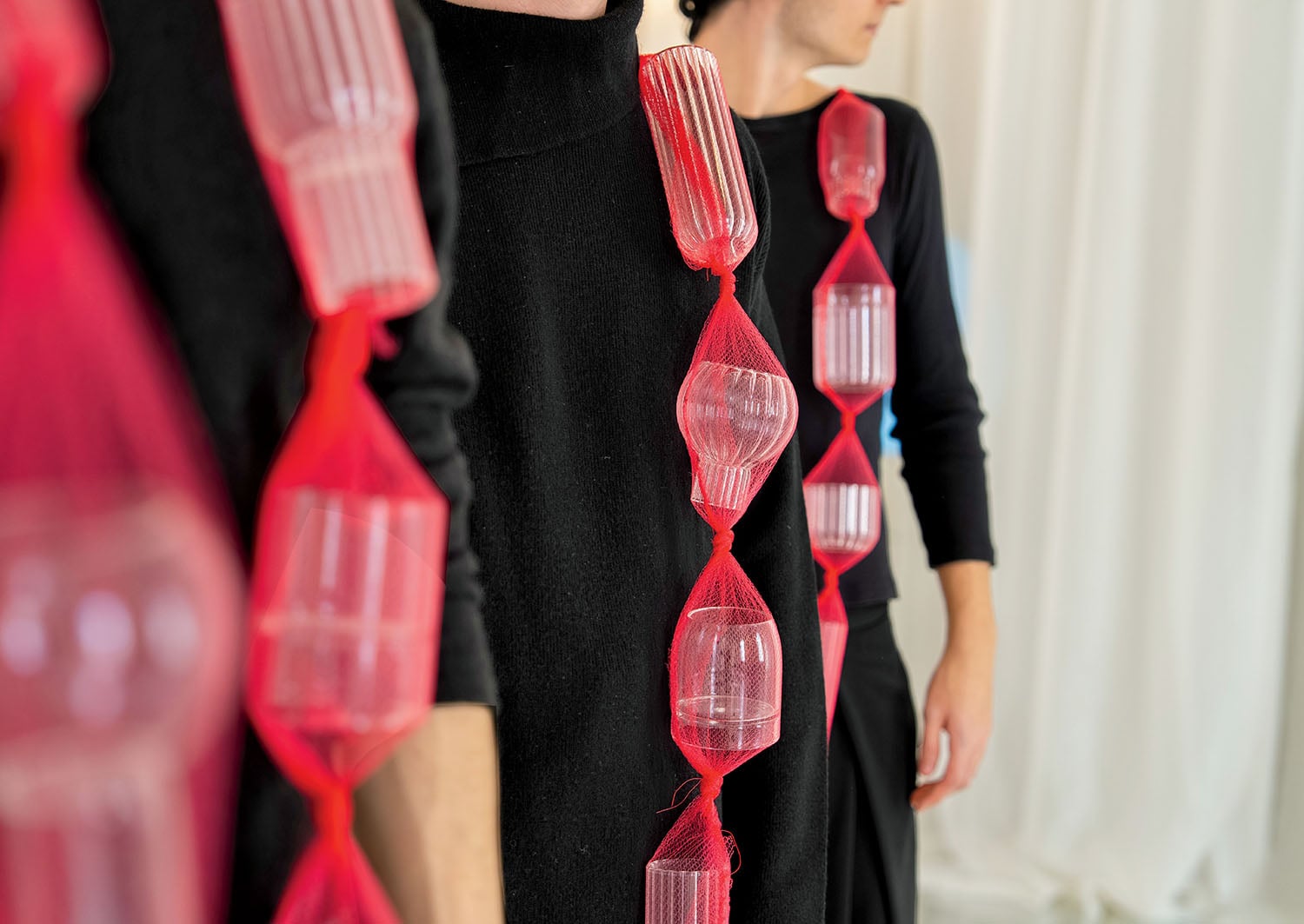 Three people wearing black outfits with red netting that holds clear plastic containers, standing in a minimalist, light-colored room.