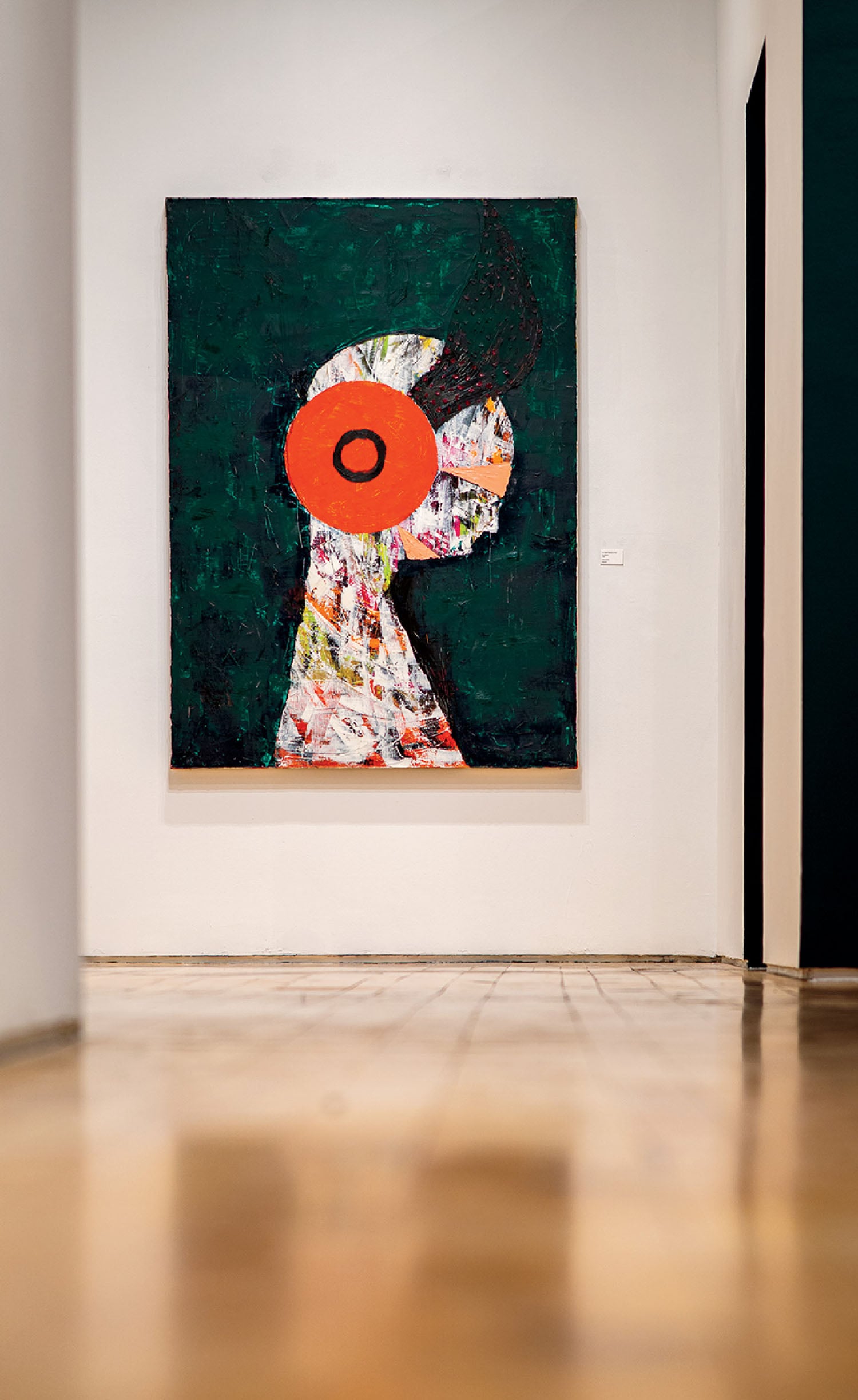 A colorful abstract painting of a human profile with a large red circle on the head hangs on a white wall in a gallery space.
