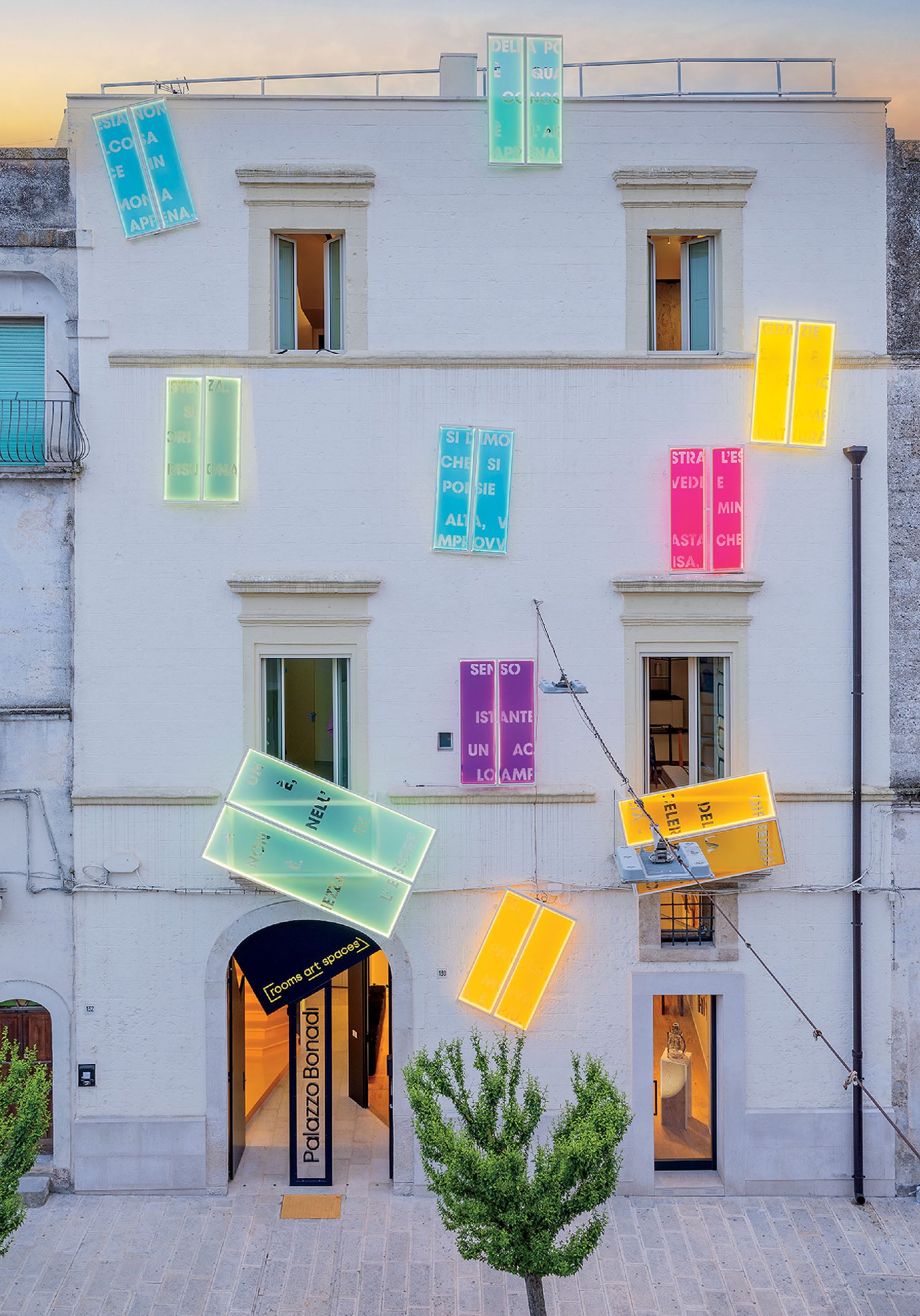 A white three-story building with brightly colored, illuminated signs and panels attached to its facade at various angles, seen from the street level.