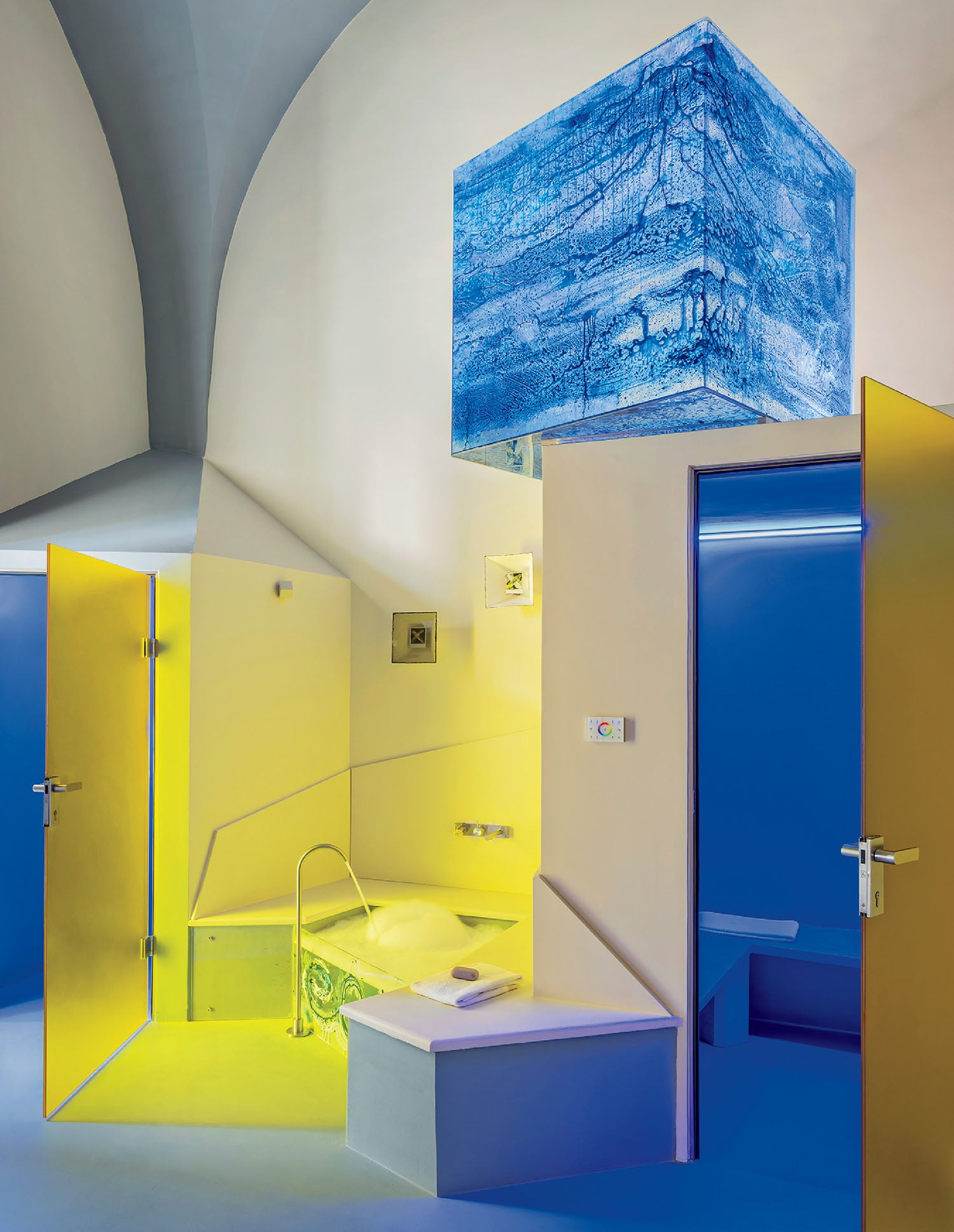 A modern bathroom with bold yellow and blue lighting, geometric walls, a sunken bathtub, and a suspended blue illuminated cube ceiling fixture.