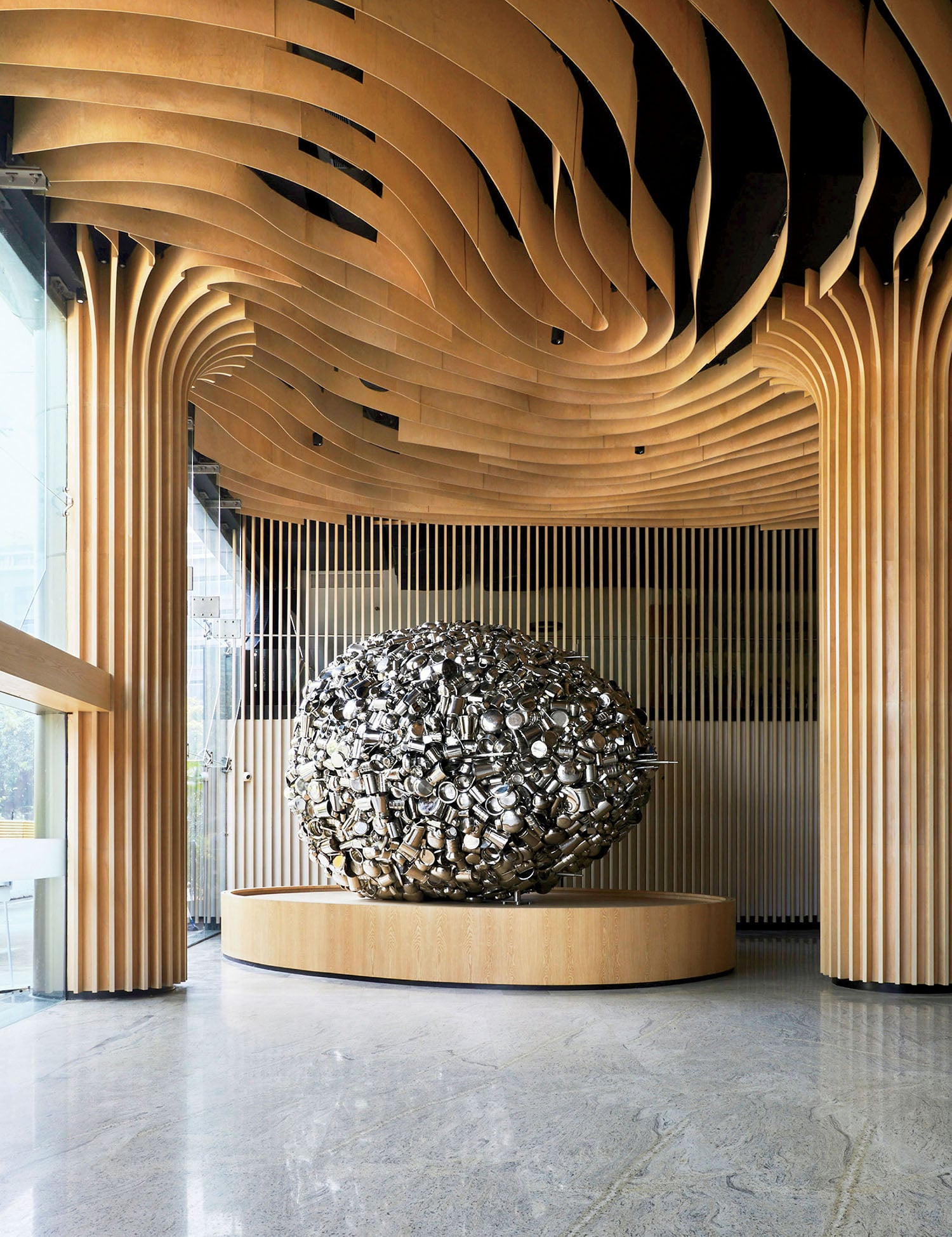 A large metallic abstract sculpture sits on a circular wooden platform beneath a ceiling with wavy wooden panels in a modern interior space.