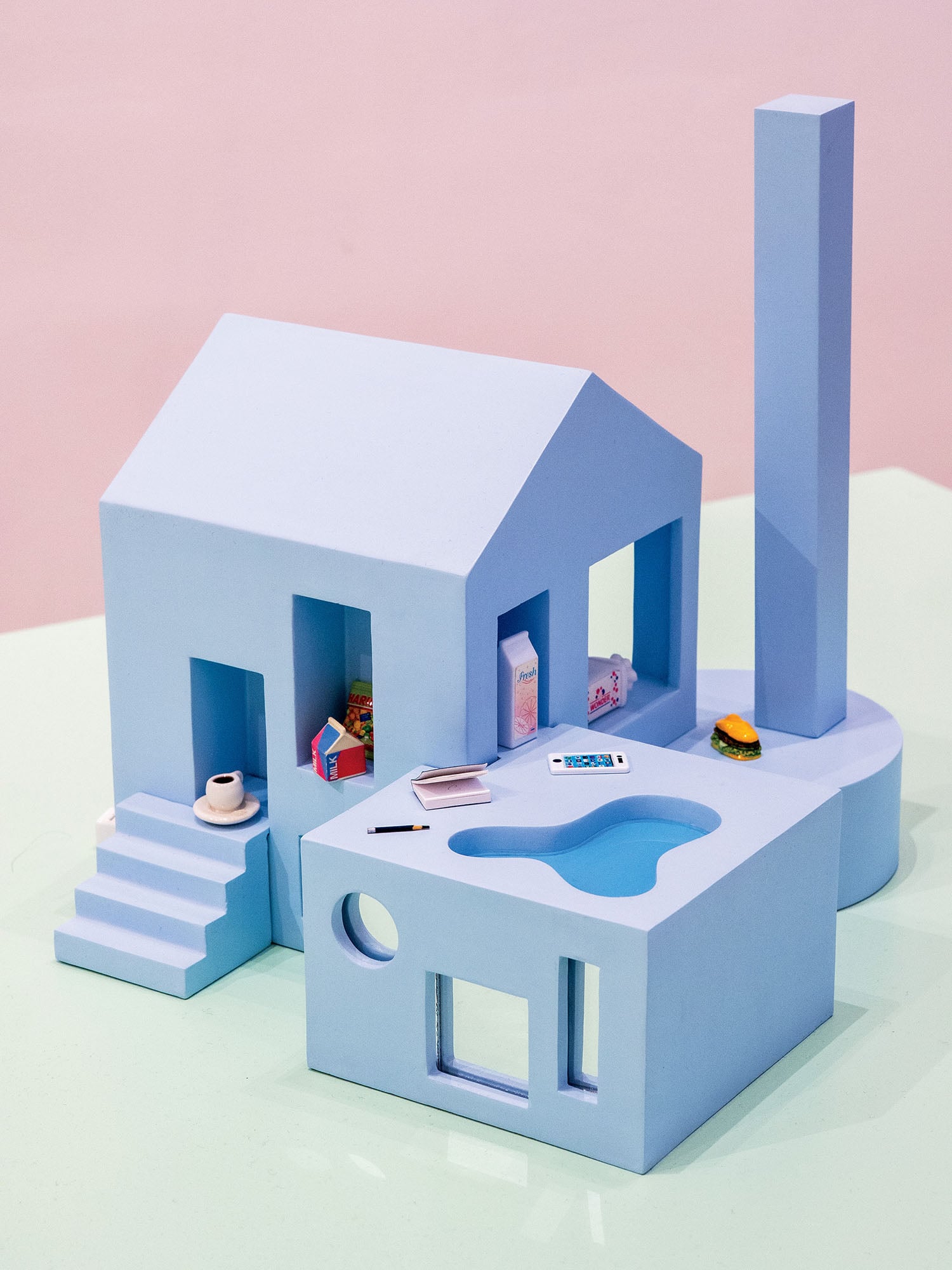 A minimalistic blue architectural model features a house, geometric structures, a pool, stairs, and small objects like food items, a notebook, and a cup placed around the model.