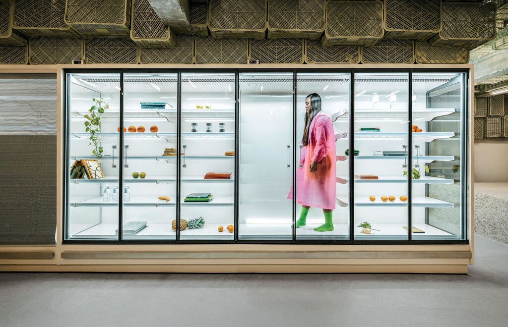 A person in a pink coat and green boots stands inside a large, mostly empty glass display fridge with scattered fruits, vegetables, books, and plants on the shelves.