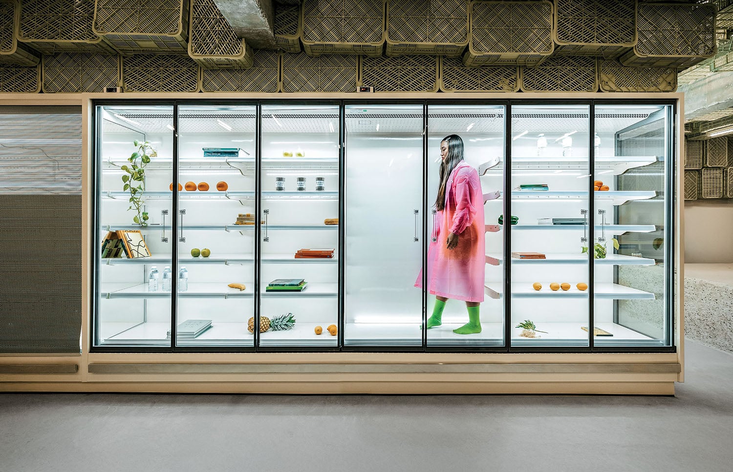 A person in a pink coat and green boots stands inside a large, mostly empty glass display fridge with scattered fruits, vegetables, books, and plants on the shelves.