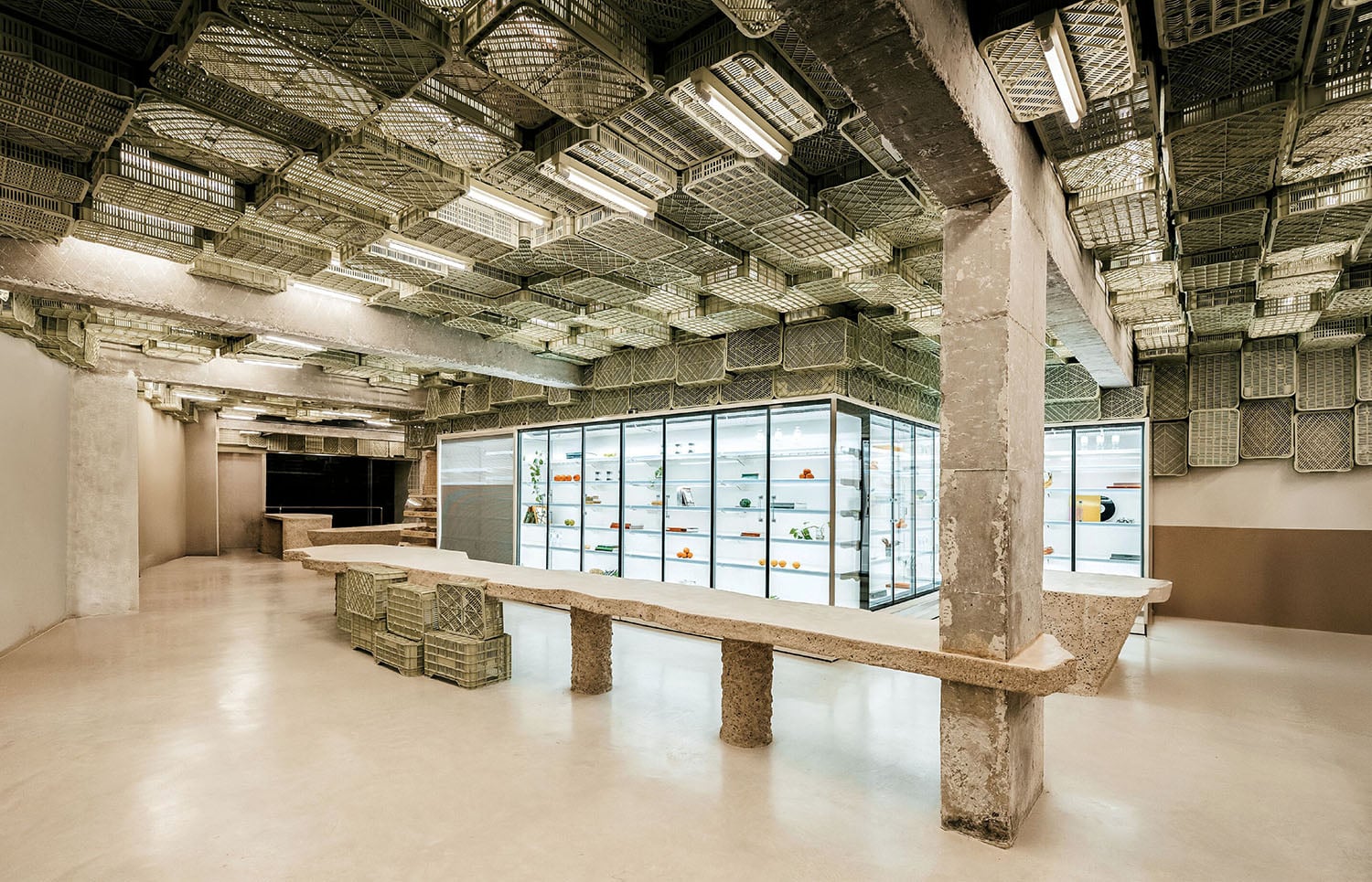 A modern, minimalist store interior with exposed concrete, a long stone table, baskets on the ceiling, and a glass display case with various items inside.