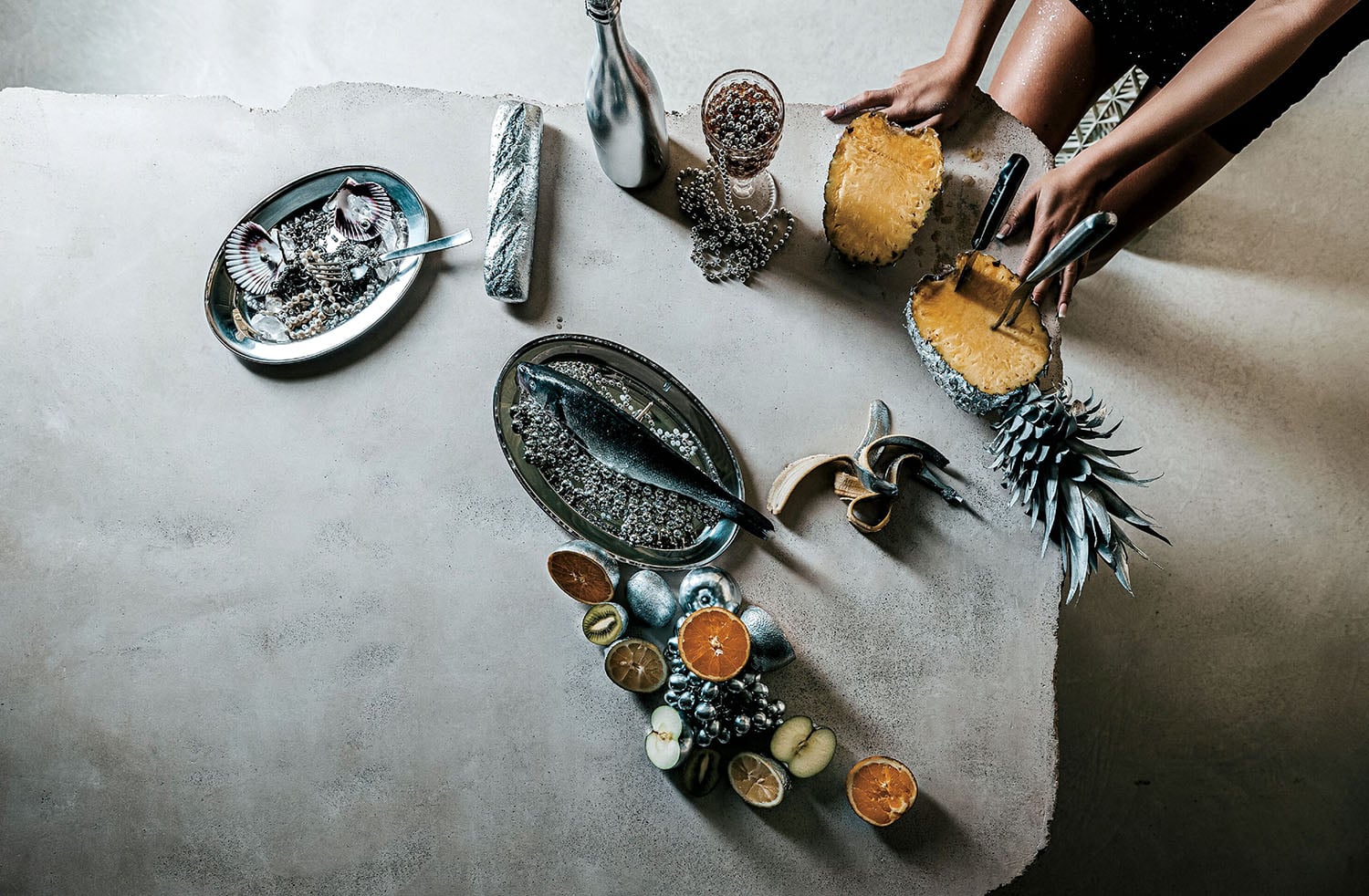 Overhead view of a table with various foods including a whole fish, citrus fruits, a wrapped item, drinks, and a person slicing a pineapple.
