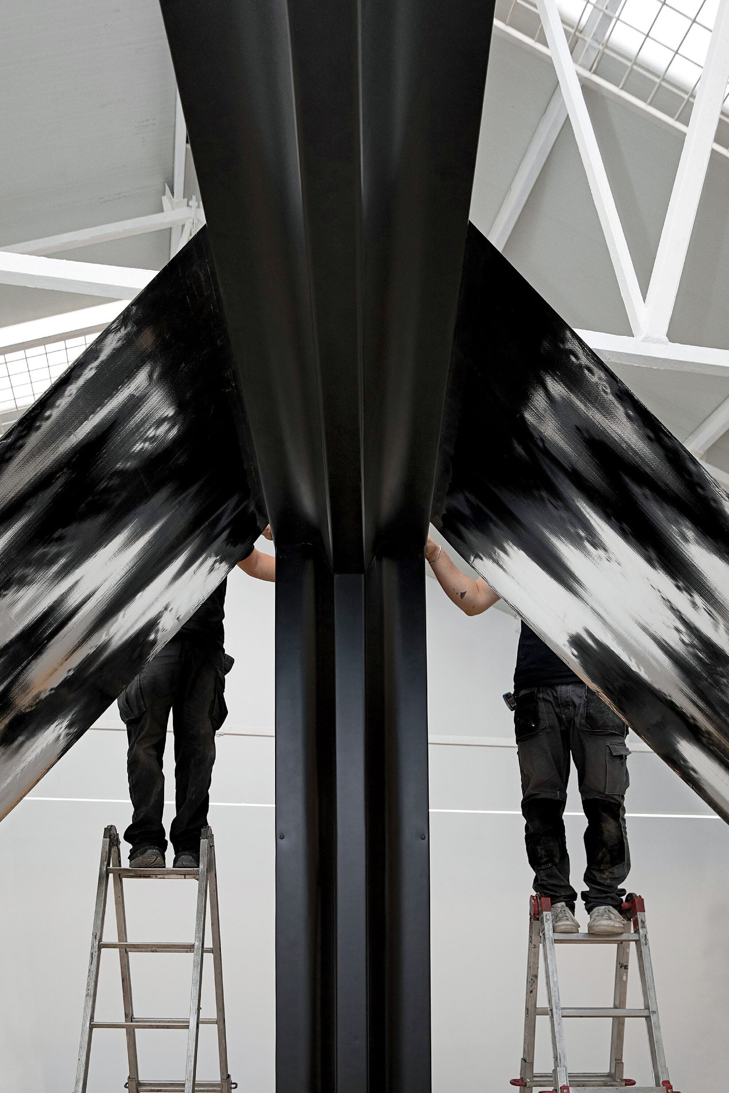 Two people stand on ladders, working on a large black and white metal structure inside a building with a high, white ceiling and industrial lighting.