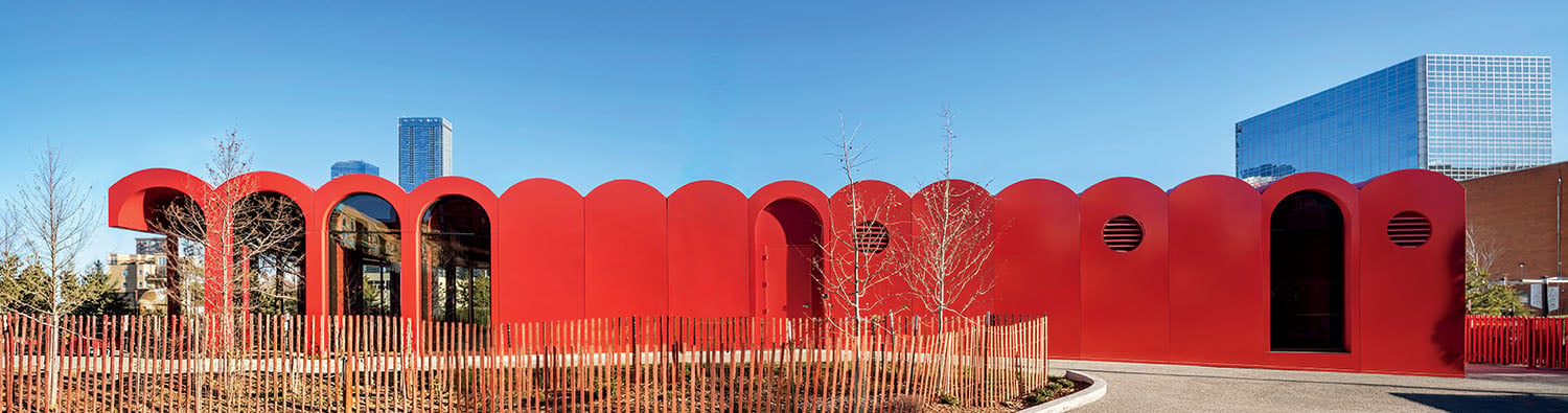 A modern building with a bright red, scalloped roofline and arched doors and windows, set against a blue sky and skyscrapers in the background.