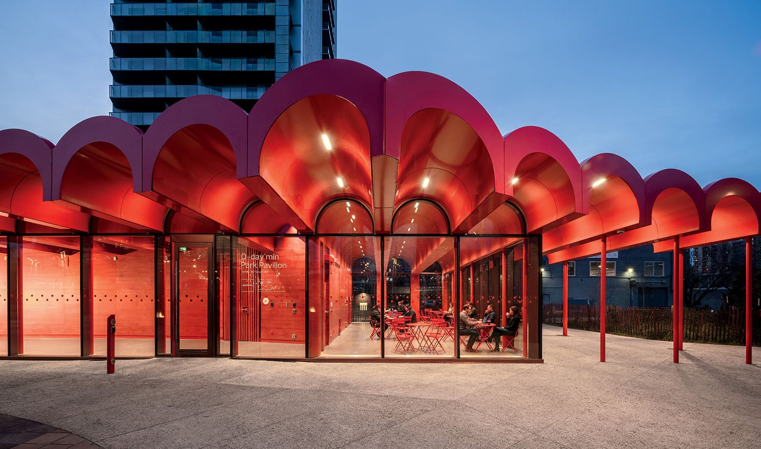 A modern building with a bright red scalloped roof, glass walls, and visible seating area inside, set against an evening sky with a high-rise building in the background.