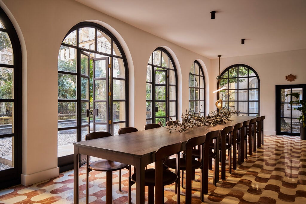 A long wooden dining table with chairs sits in a bright room with arched windows, patterned tile floor, and a contemporary branch centerpiece.
